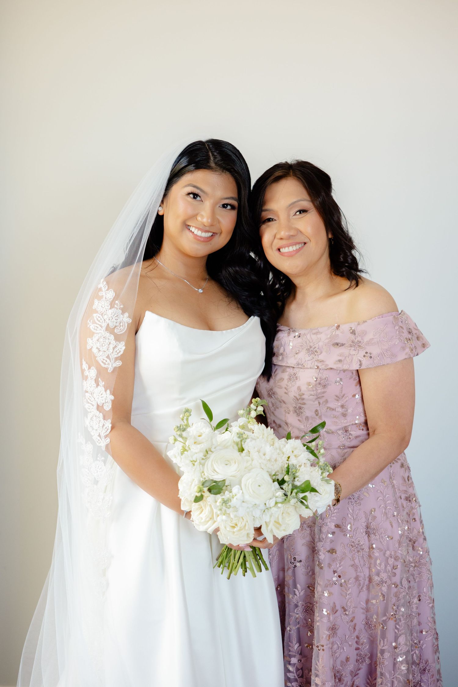 Joyful wedding portrait with bride in white gown and woman in dusty purple lace dress.
