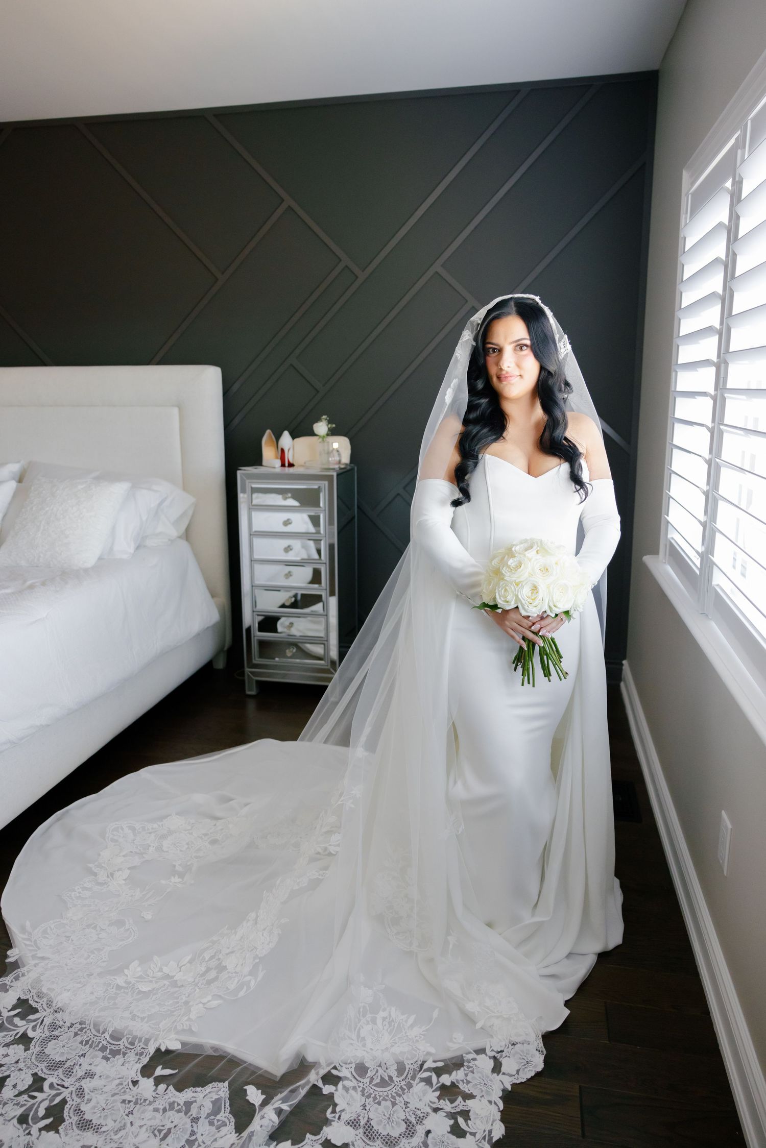 A bride in a fitted white gown with long veil holds a white bouquet in a modern bedroom setting.
