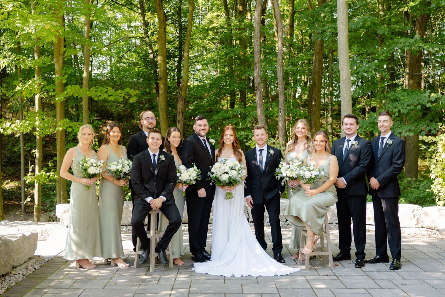 Wedding party poses together outdoors in formal attire against lush green forest backdrop.