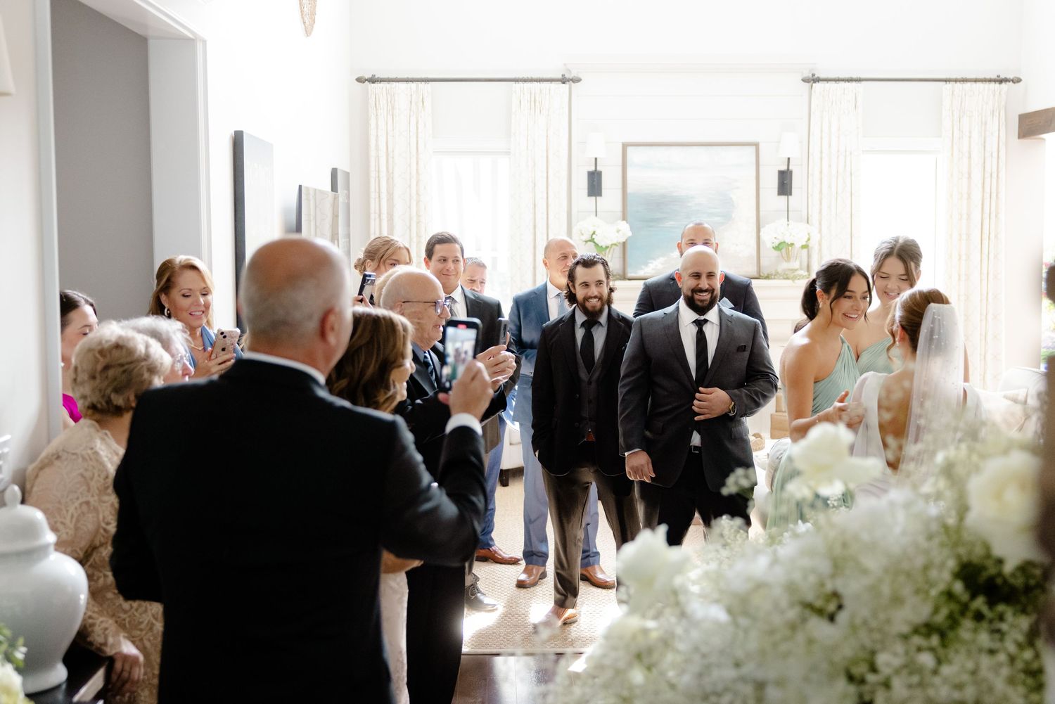 Wedding ceremony entrance with guests gathered in bright white room filled with floral arrangements.