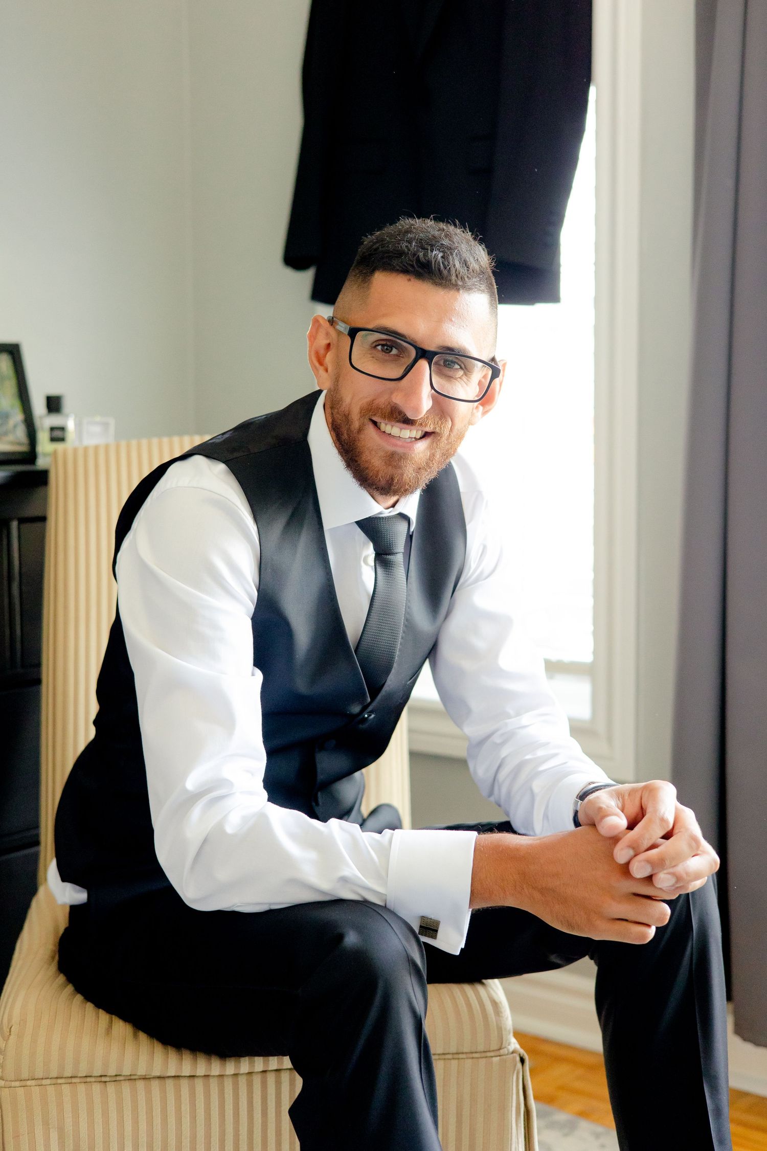 Formal portrait of someone wearing a vest and tie sitting on a beige chair.
