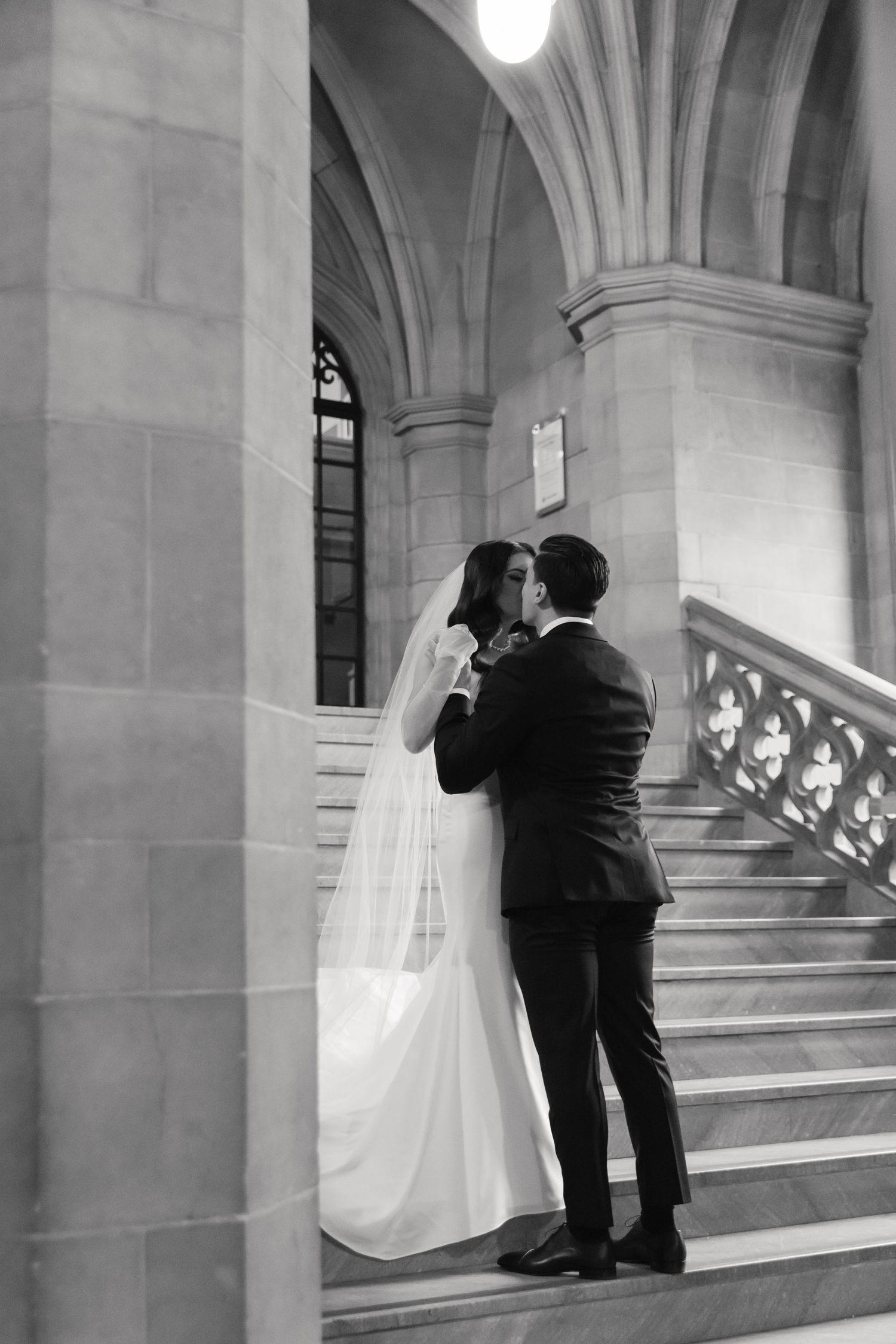 A romantic wedding moment captured in black and white on an elegant stone staircase with gothic architectural details.