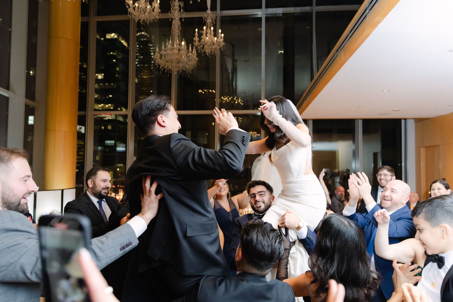 Bride and groom being lifted on chairs during traditional wedding celebration dance.
