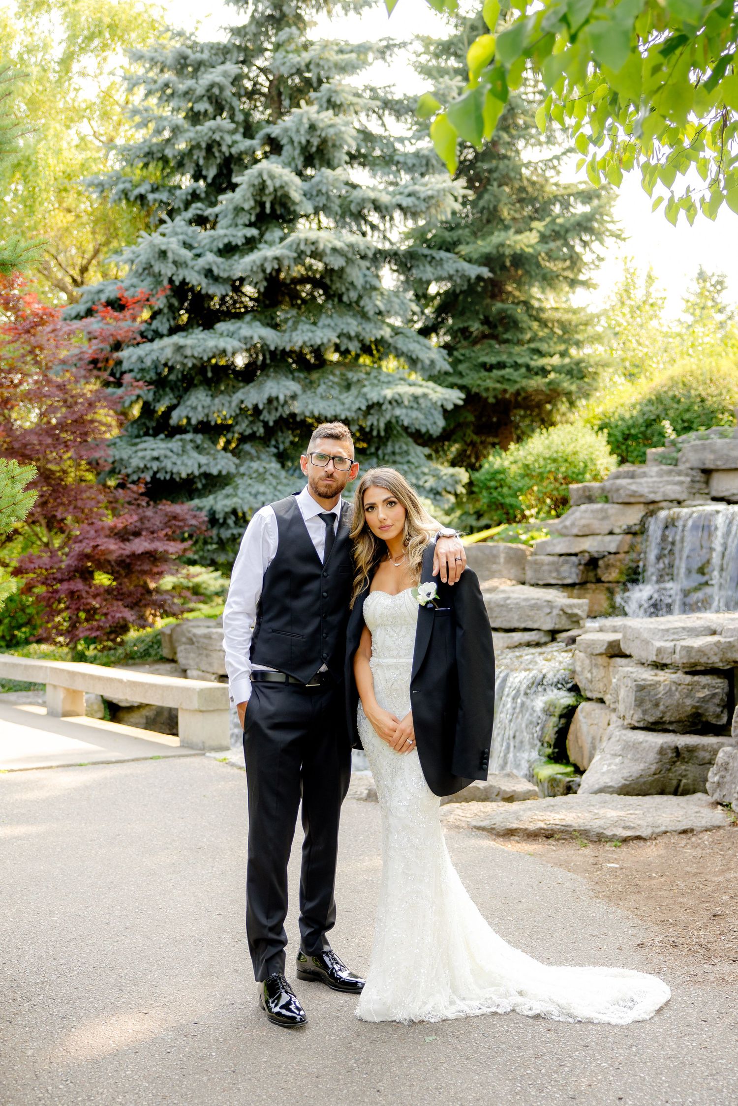 A wedding couple poses formally in front of a garden waterfall with evergreen trees in the background.