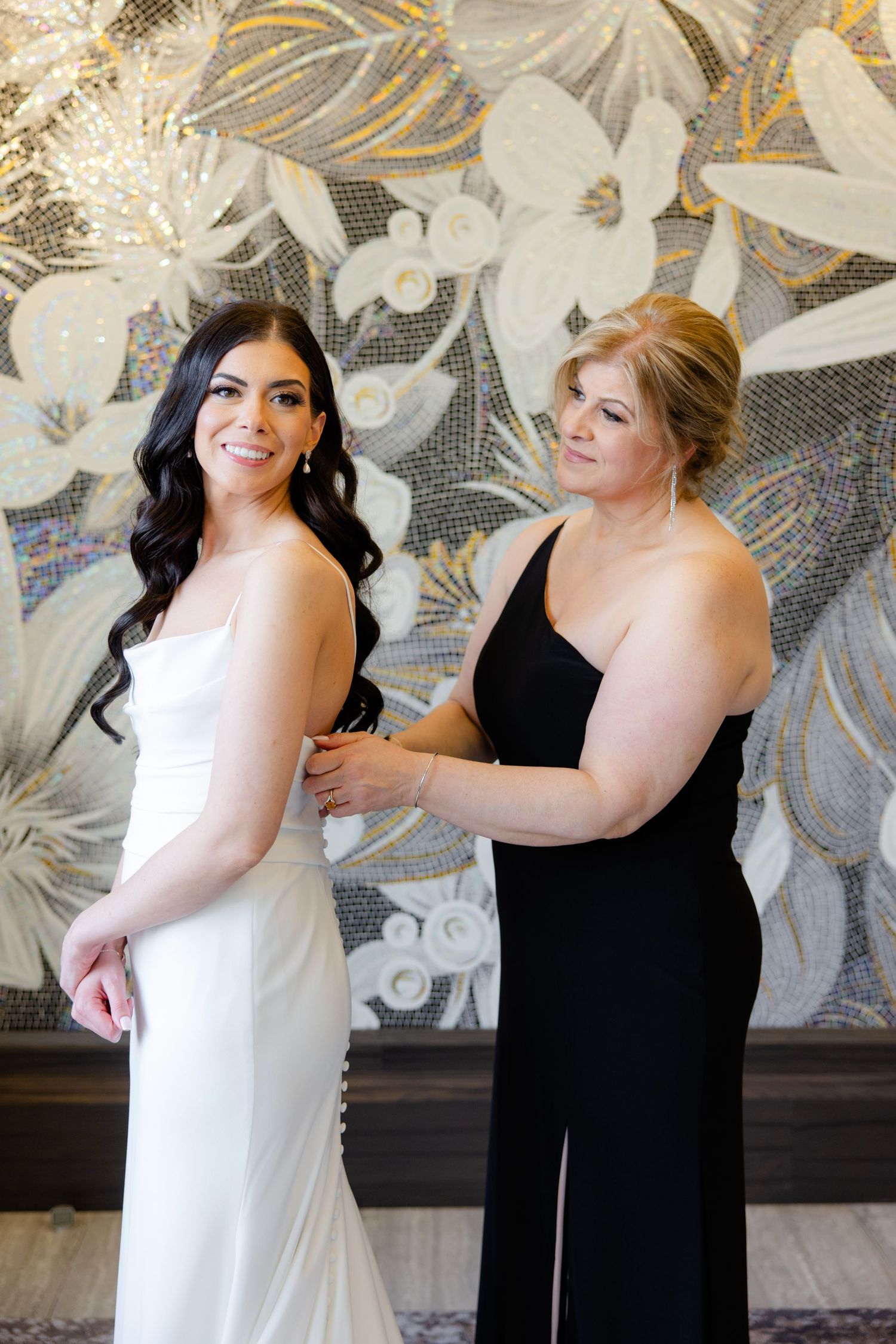 Two people in formal attire pose against an elegant floral wall during wedding preparations.