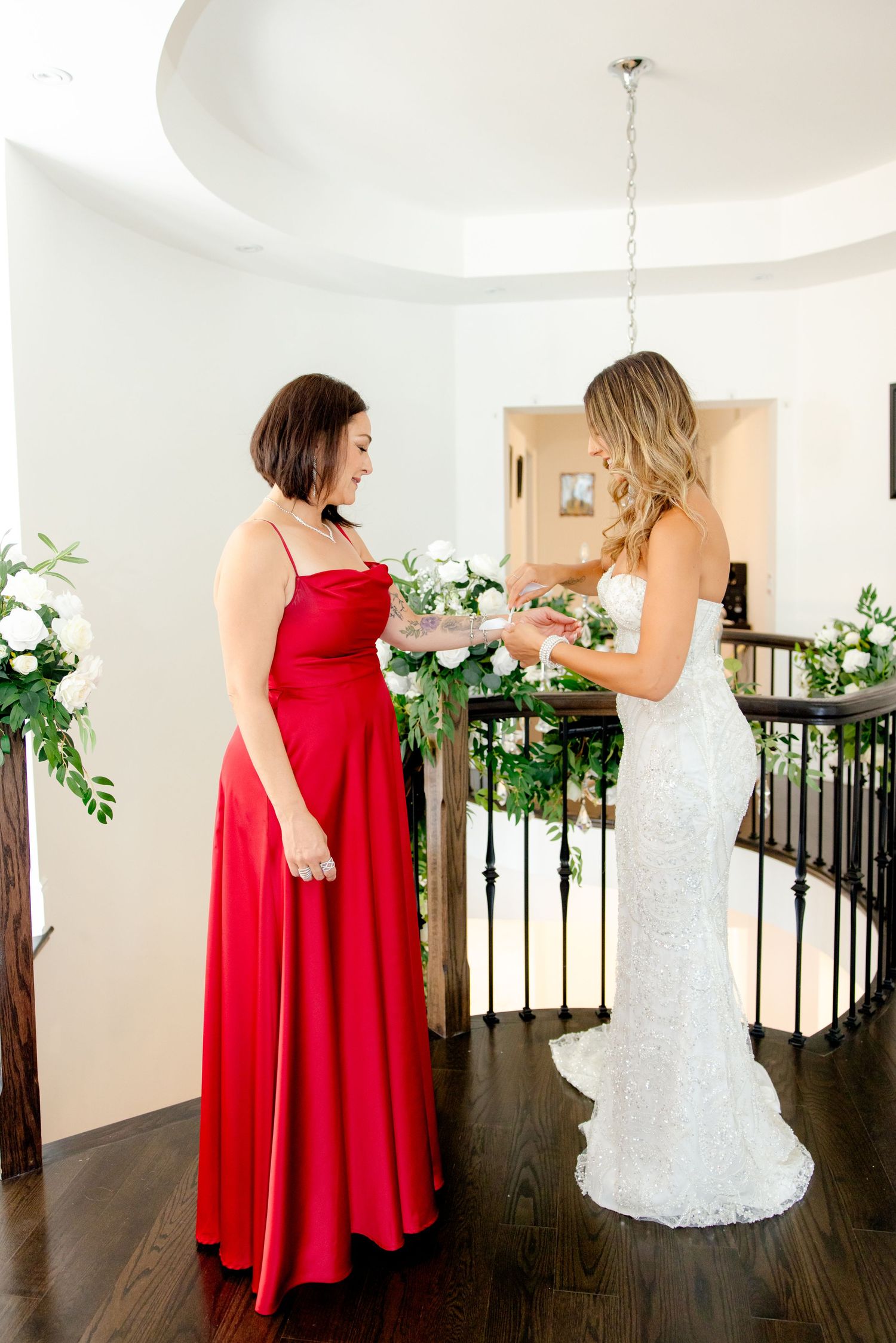 Bride in lace gown and attendant in red dress sharing moment with bouquet.