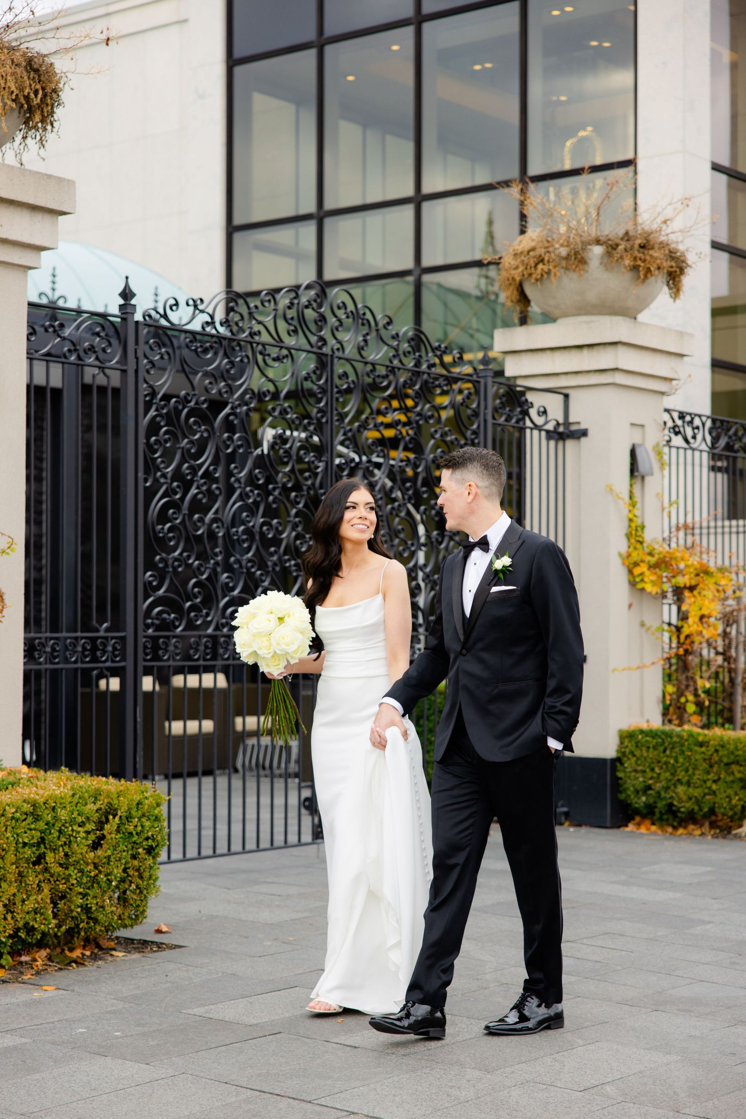 Bride holding white bouquet and groom stand together by ornate black iron gates and modern architecture.