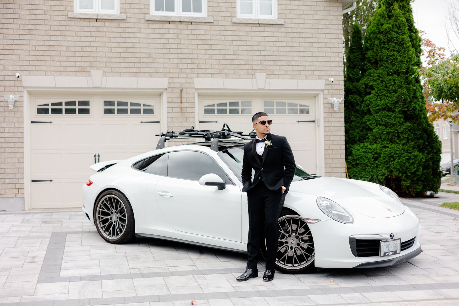 Wedding day portrait featuring a luxury white Porsche and someone in formal attire by a brick garage.