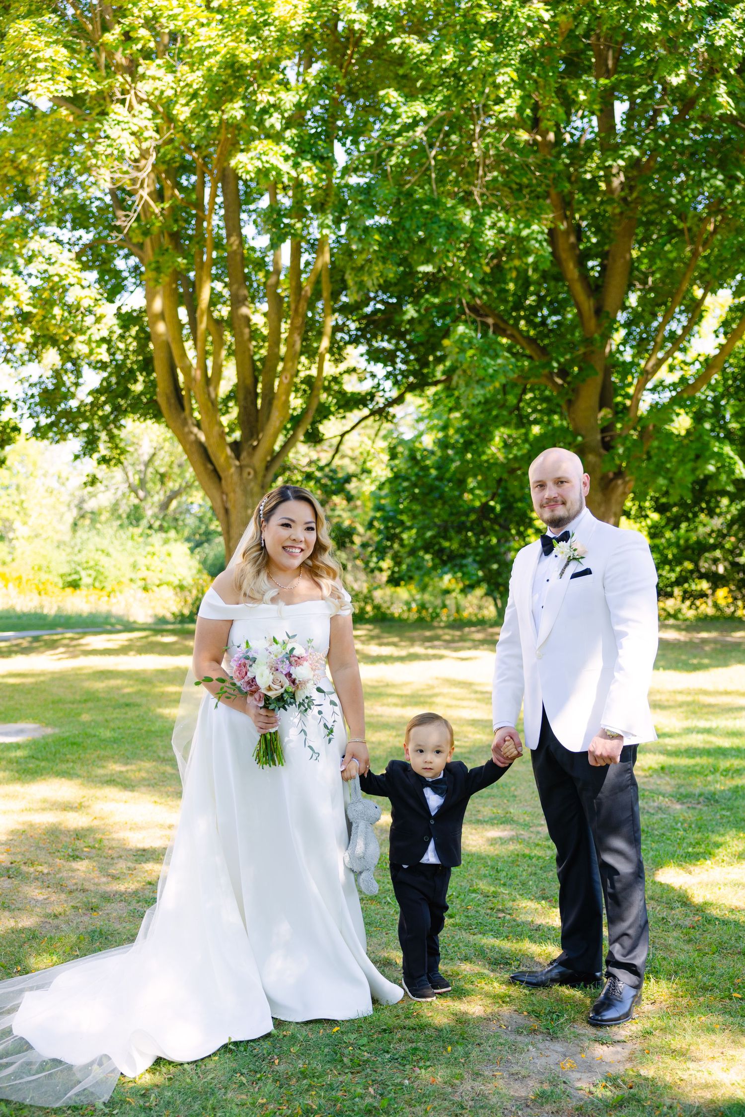 Summer wedding photo with bride in white gown alongside groom and child on grassy lawn.