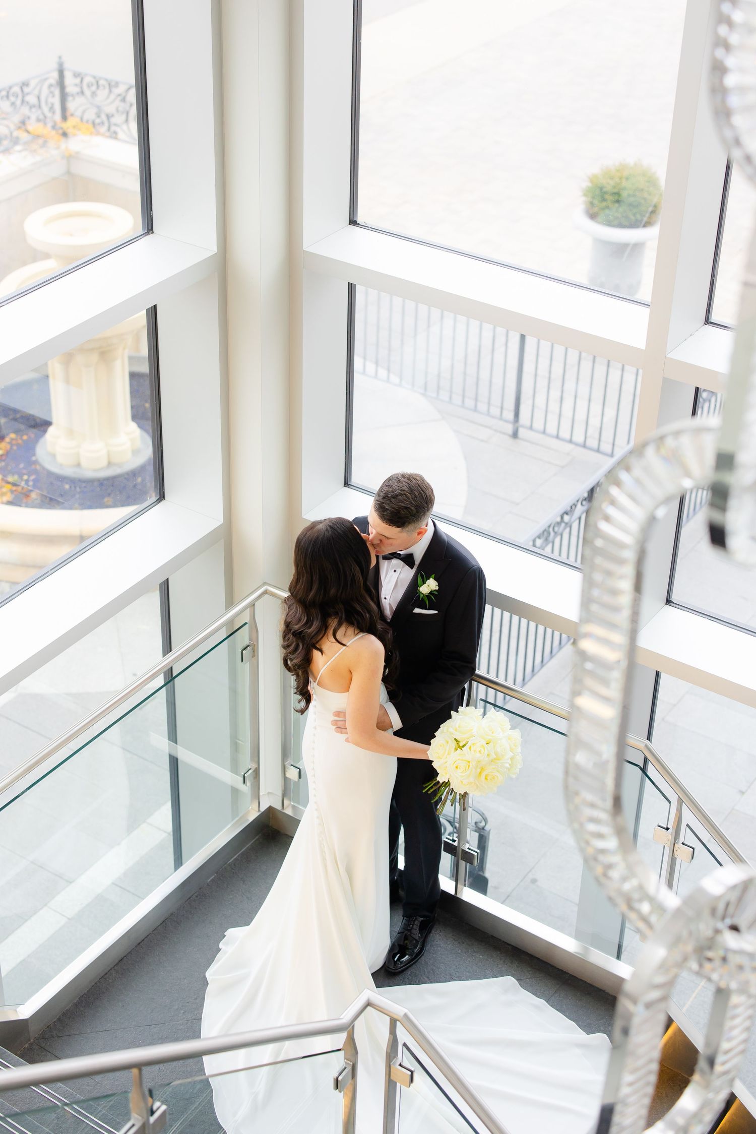 Romantic moment captured on modern staircase with crystal chandelier and natural light.