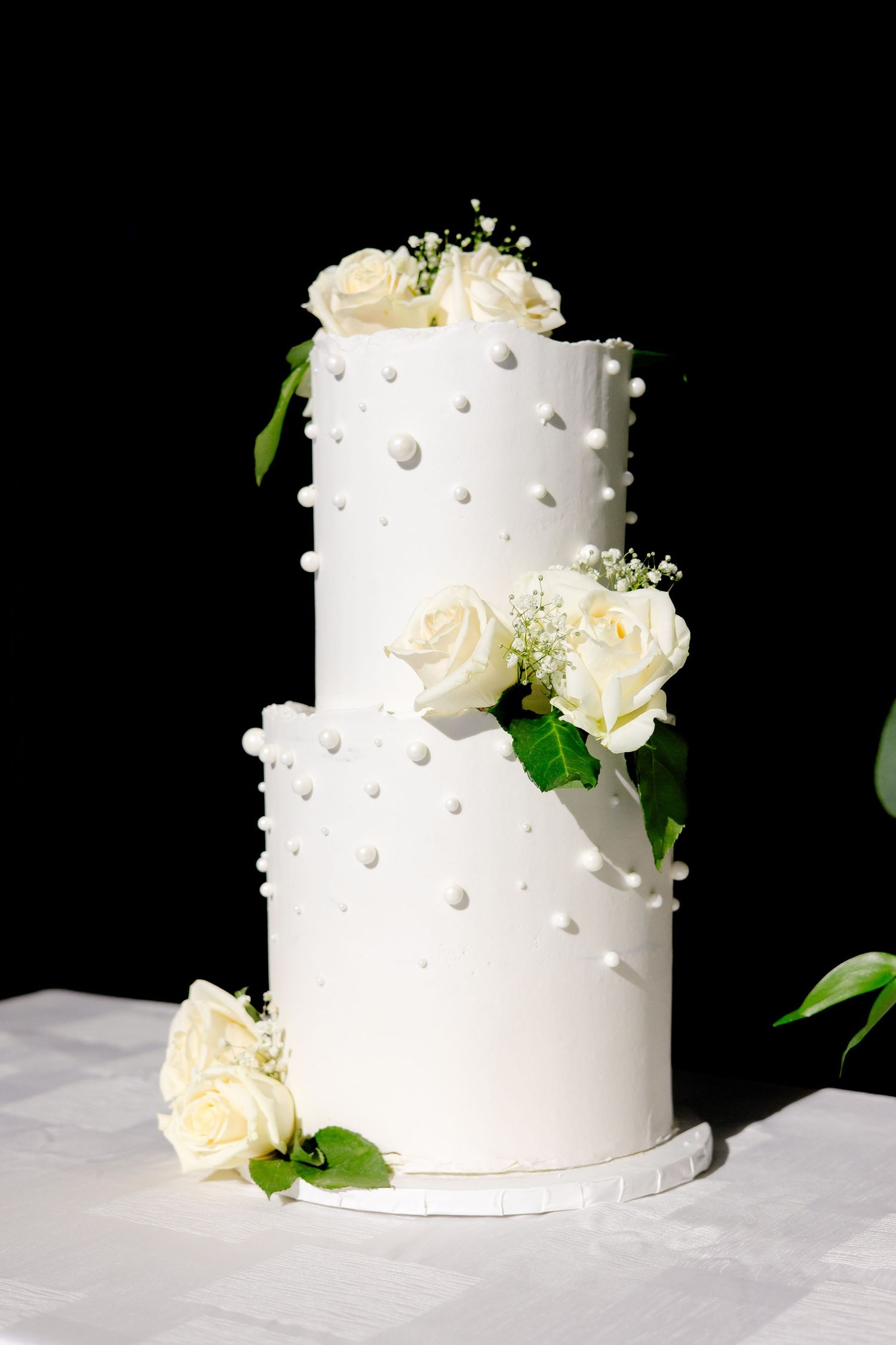 Two-tiered white wedding cake decorated with pearls and cream roses against black background.