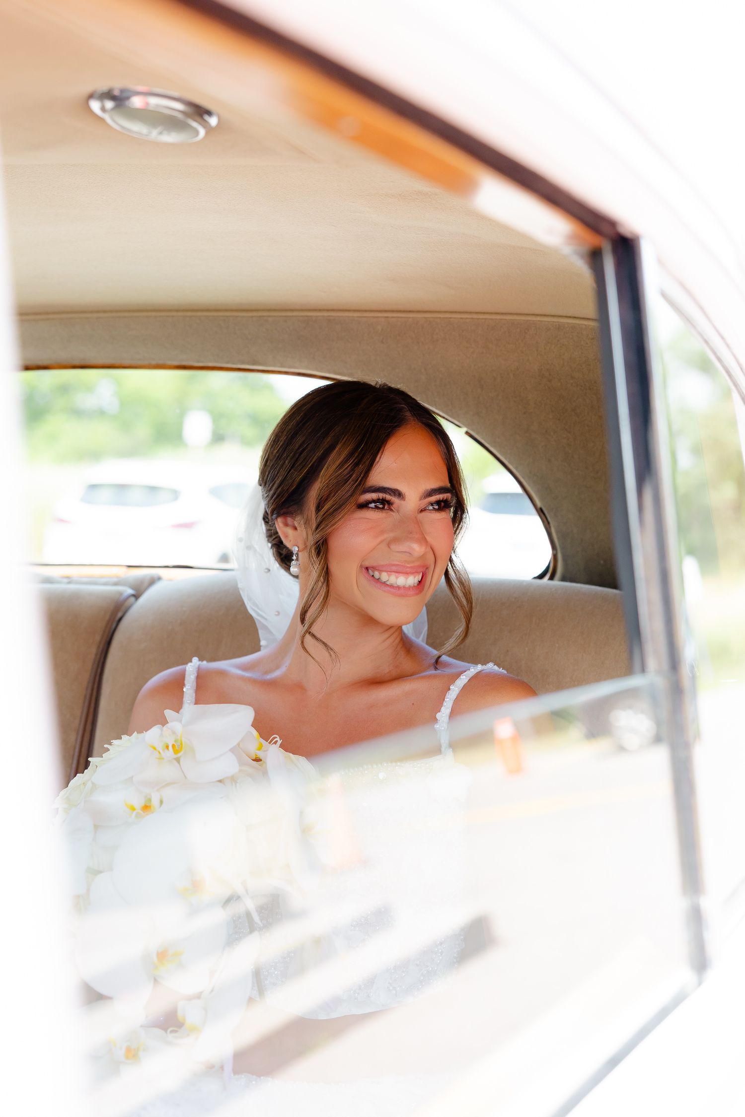 Smiling bride sitting in classic car with beige interior on her wedding day, viewed through car window.