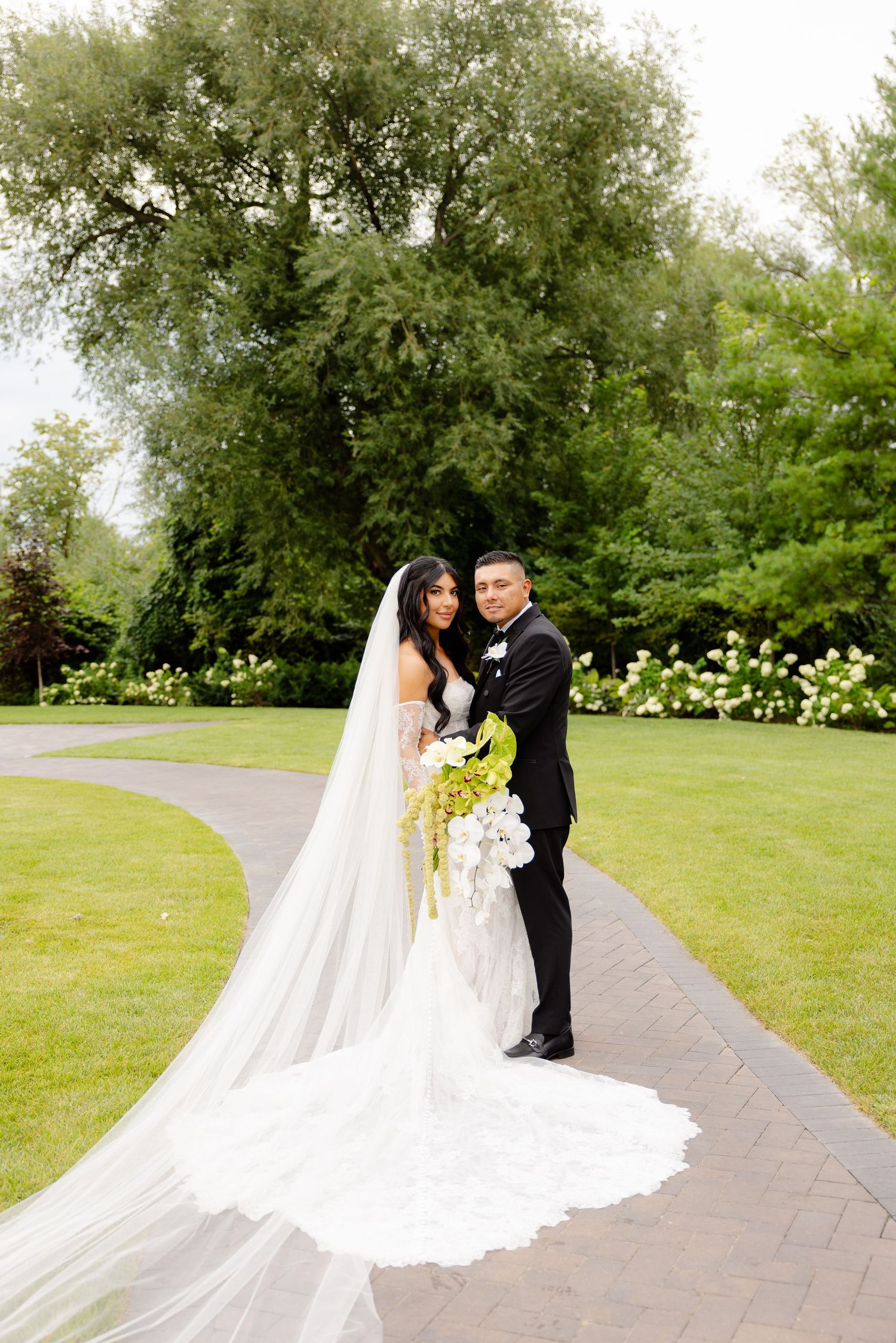 Newlyweds share intimate moments on a stone pathway surrounded by lush green landscaping and gardens.