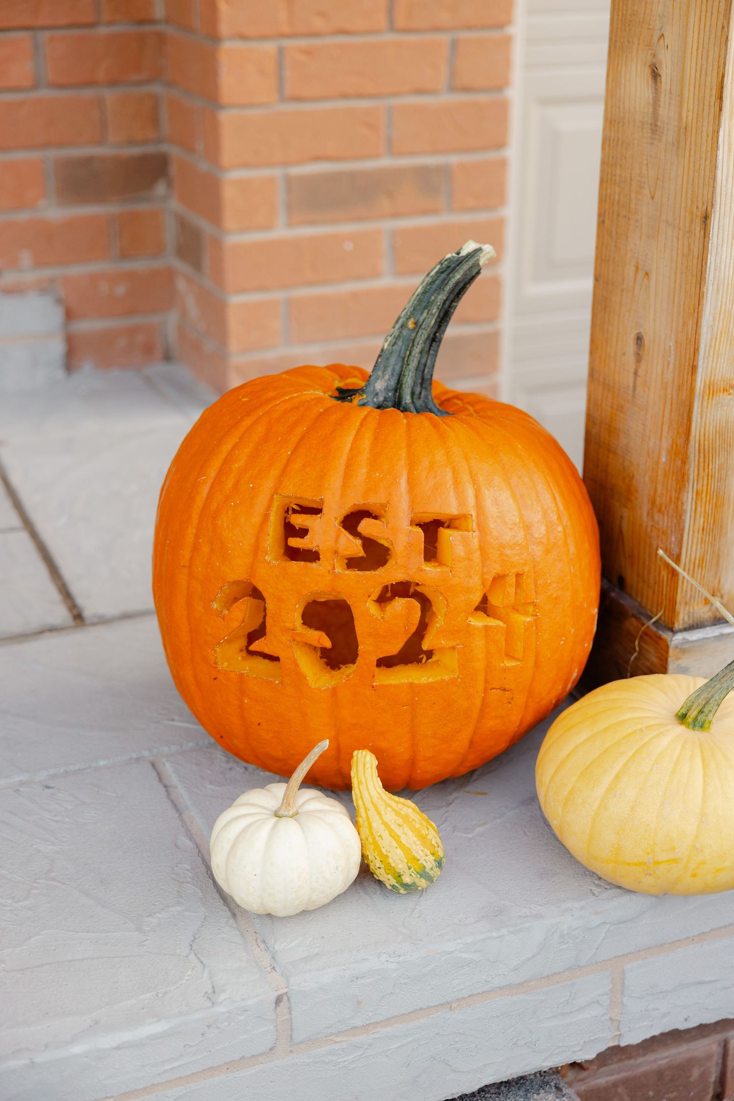 Orange pumpkin carved with 'EST 2924' sits on stone step with smaller gourds, brick wall in background.