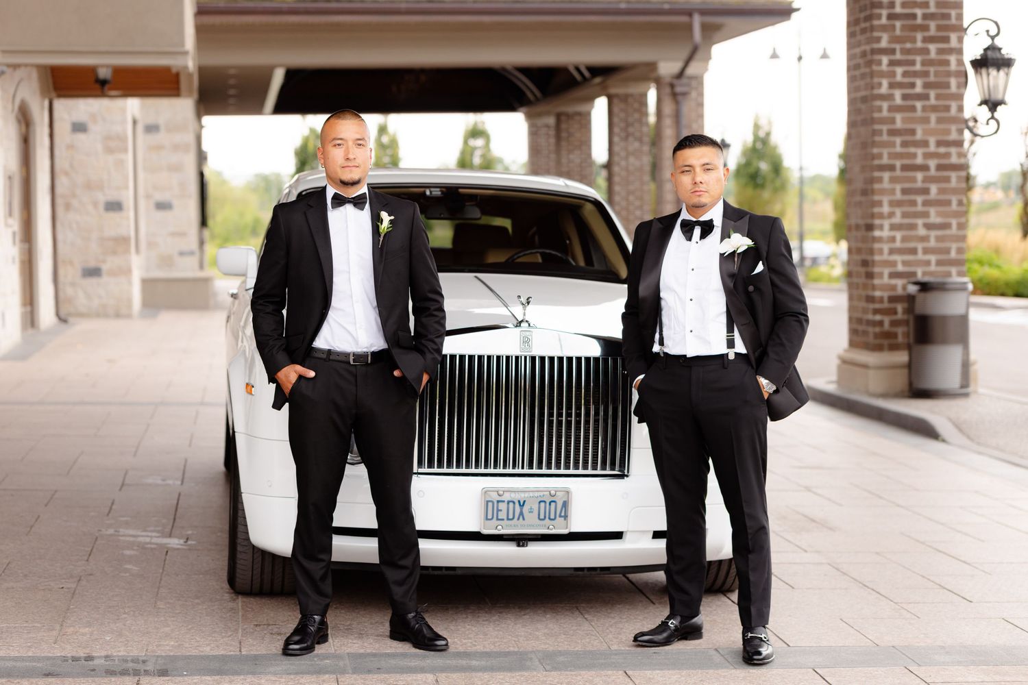 Three formal tuxedo-wearing people stand next to a black luxury car under a covered driveway.