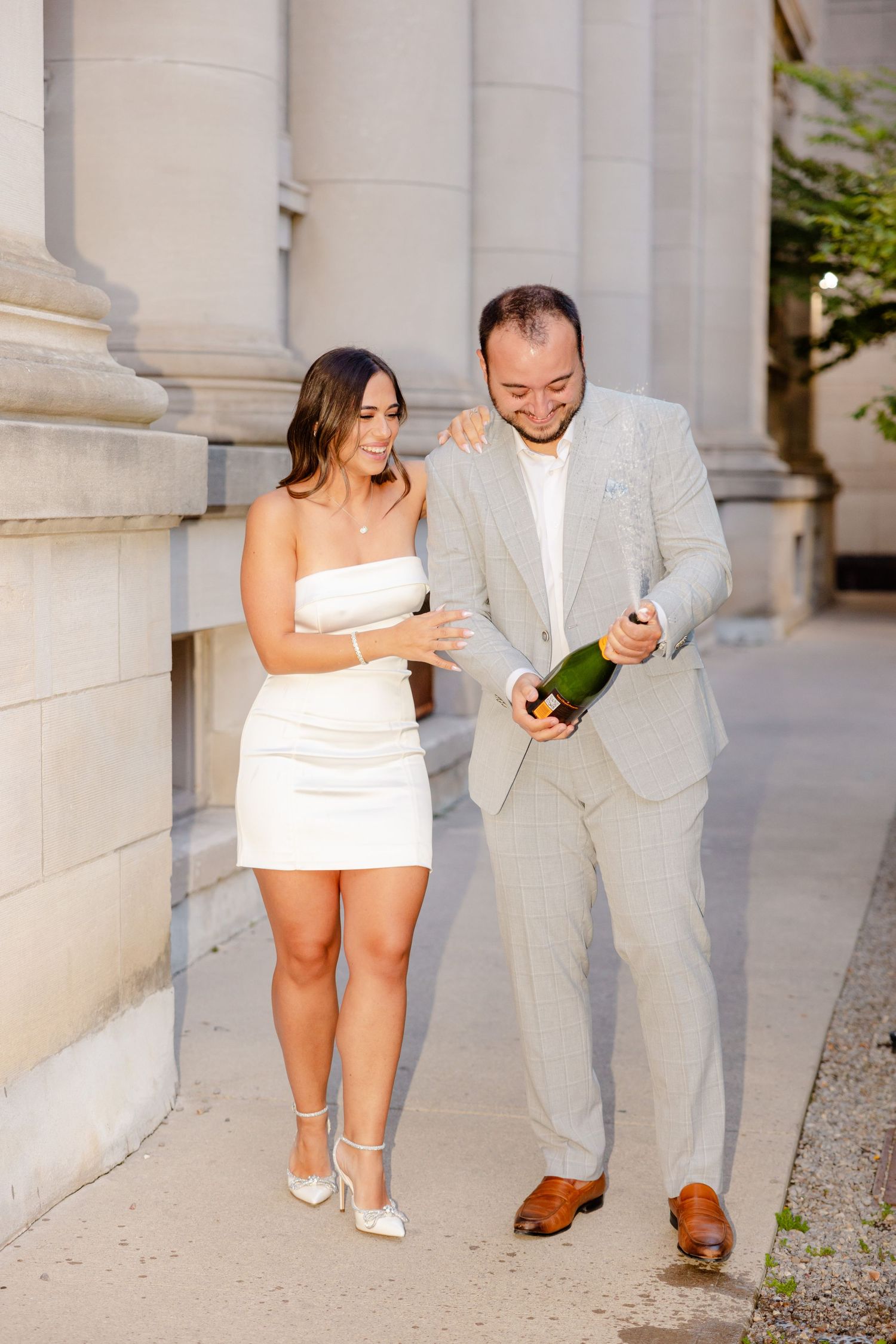 A couple shares a joyful moment with champagne while standing between elegant stone pillars.