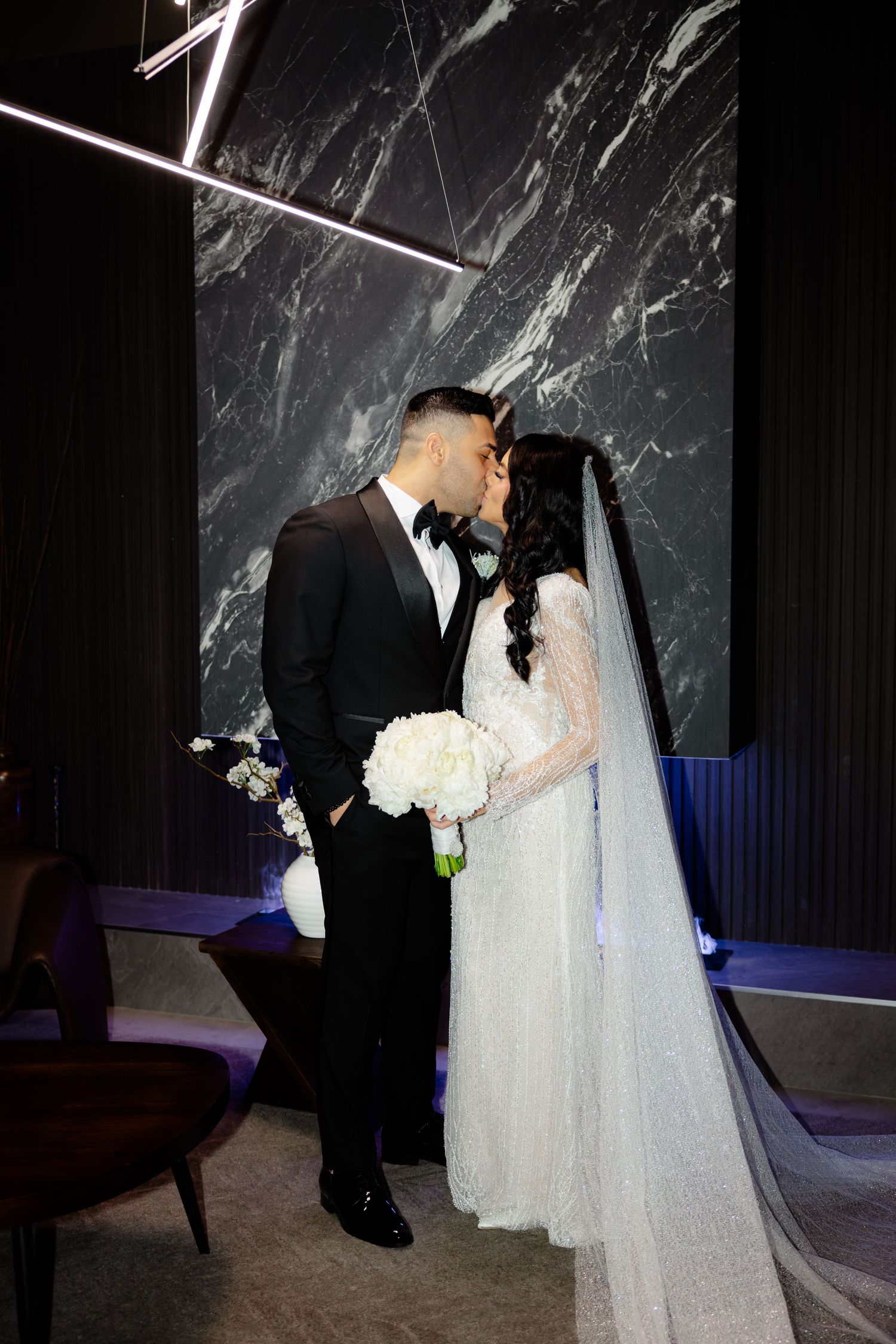 Intimate wedding portrait against marble wall with modern geometric lighting installation above.