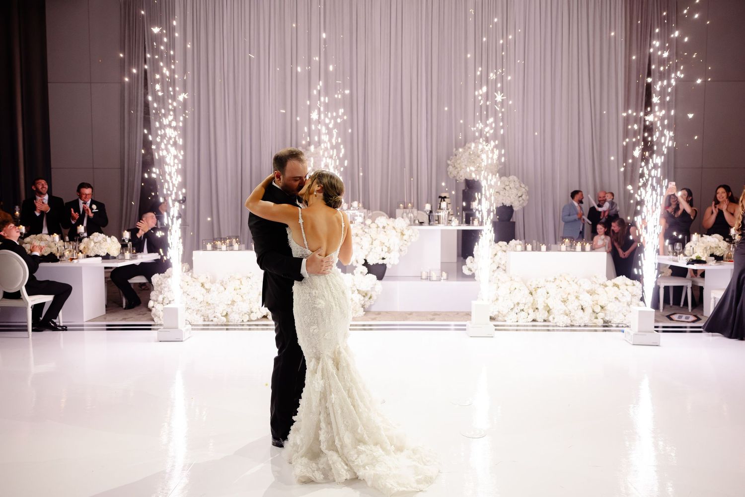 Dancing newlyweds share their first dance in an elegant white ballroom with sparklers creating a magical ambiance.