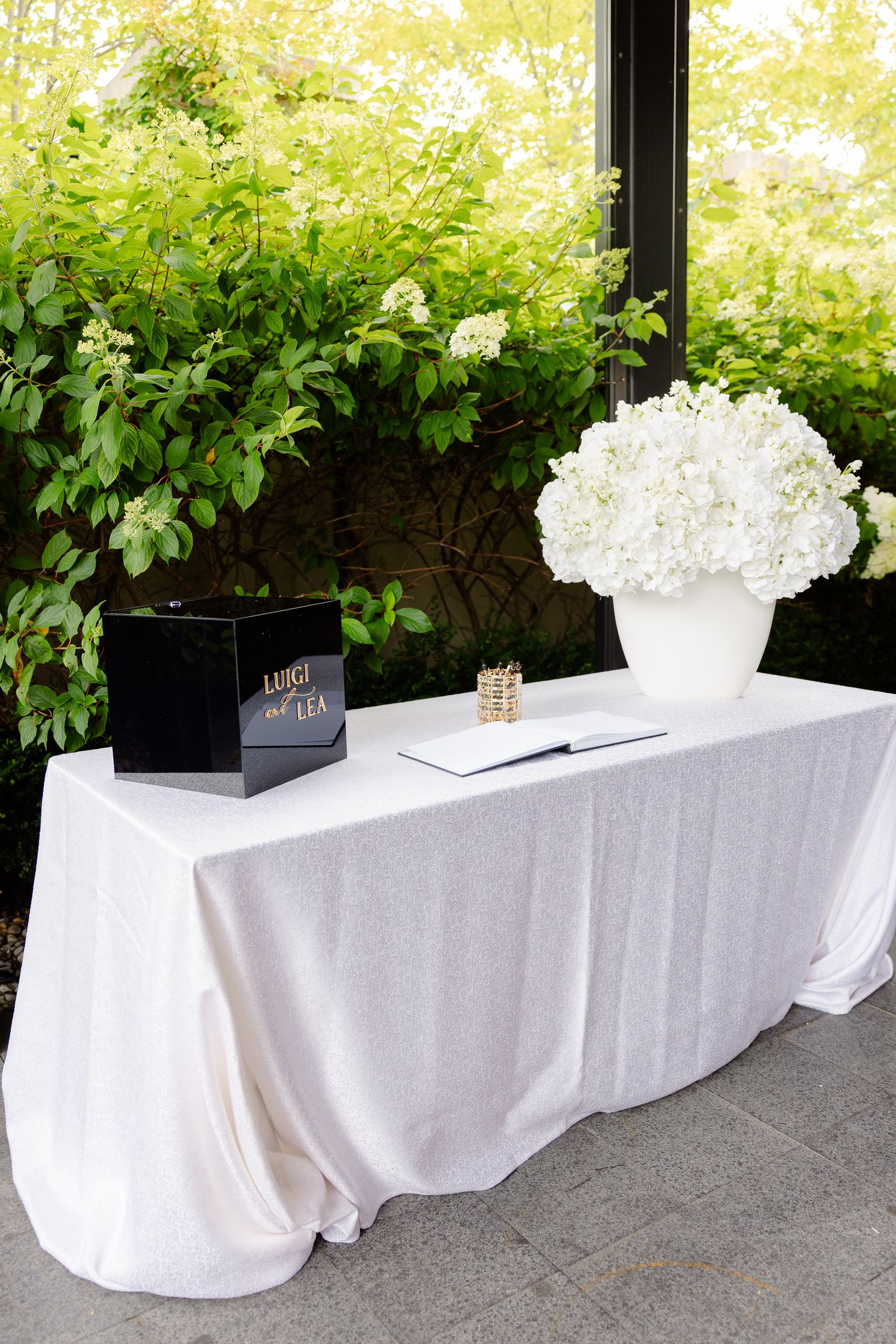 Wedding welcome table with white hydrangea centerpiece, tablecloth and black card box against greenery backdrop.