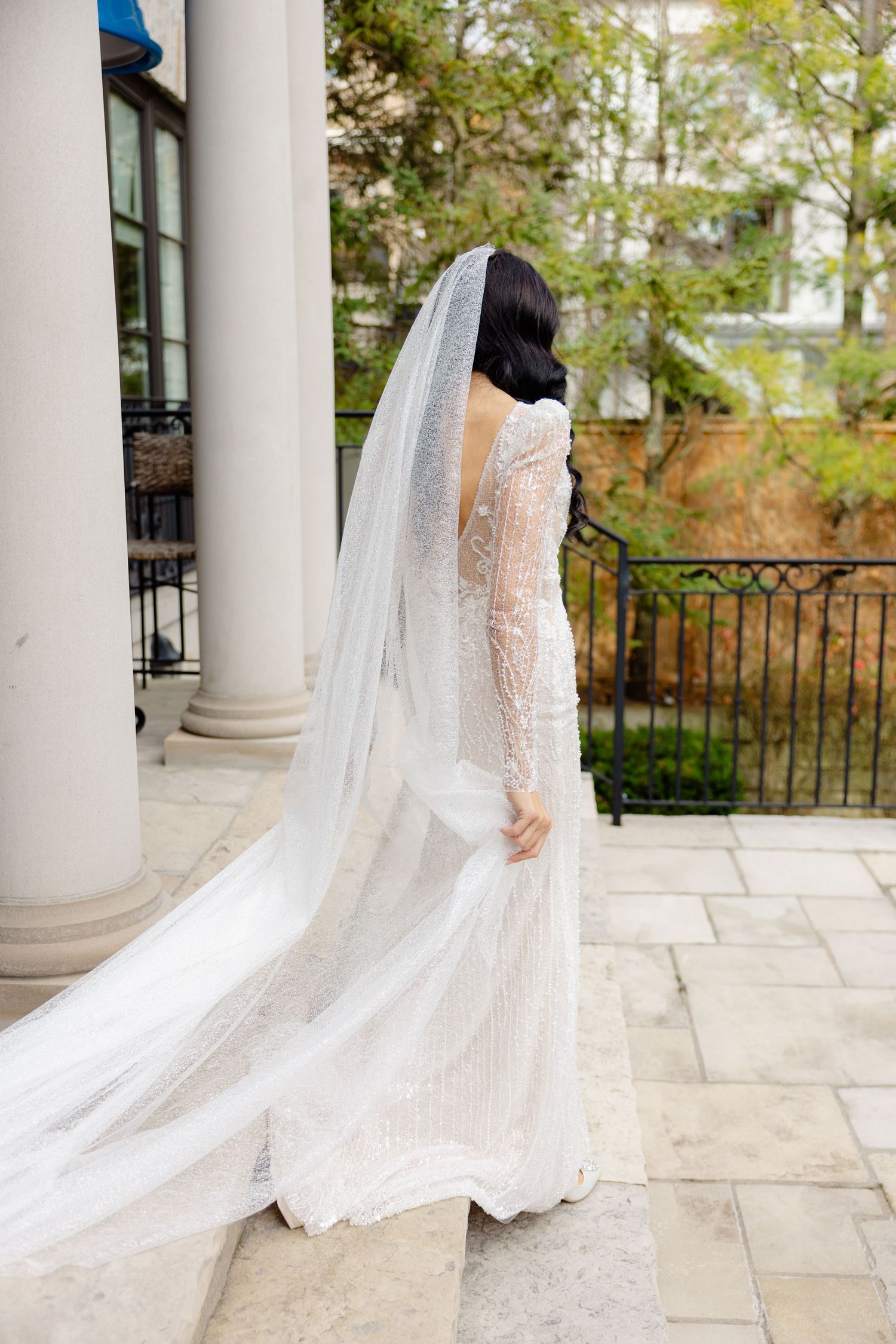 Side view of bridal gown with long cathedral veil on stone patio with white columns.