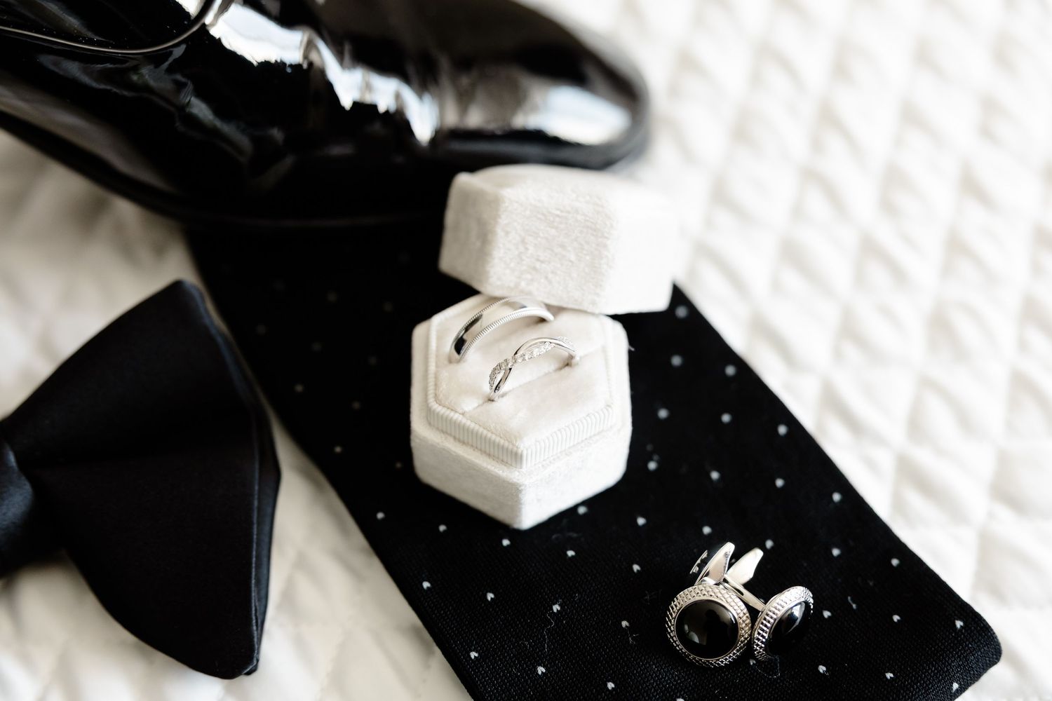 Wedding accessories including black bow tie, hexagonal ring box, and silver cufflinks arranged on polka dot fabric.