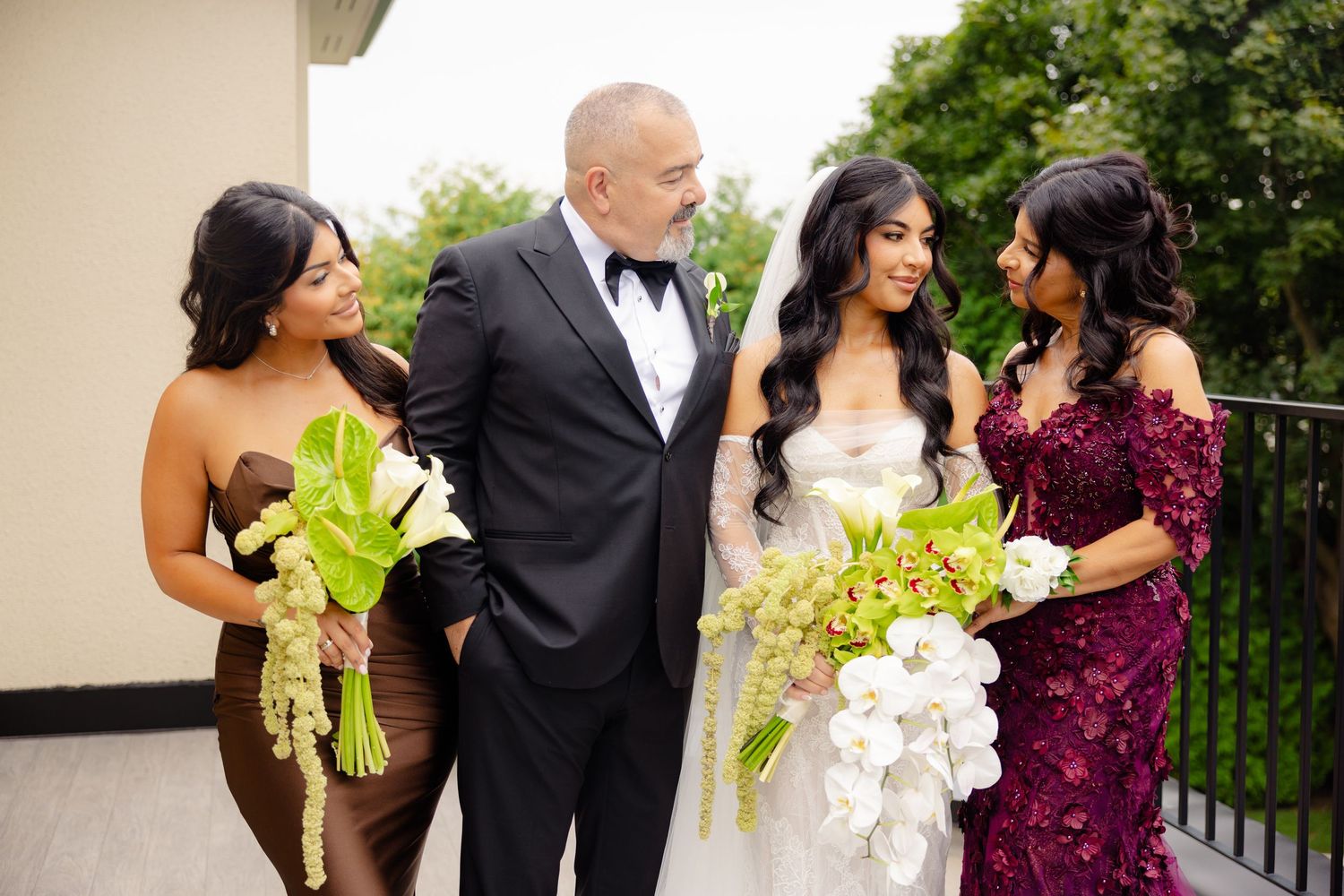 Wedding party laughs together while posing along outdoor railing with bright green and burgundy floral bouquets.
