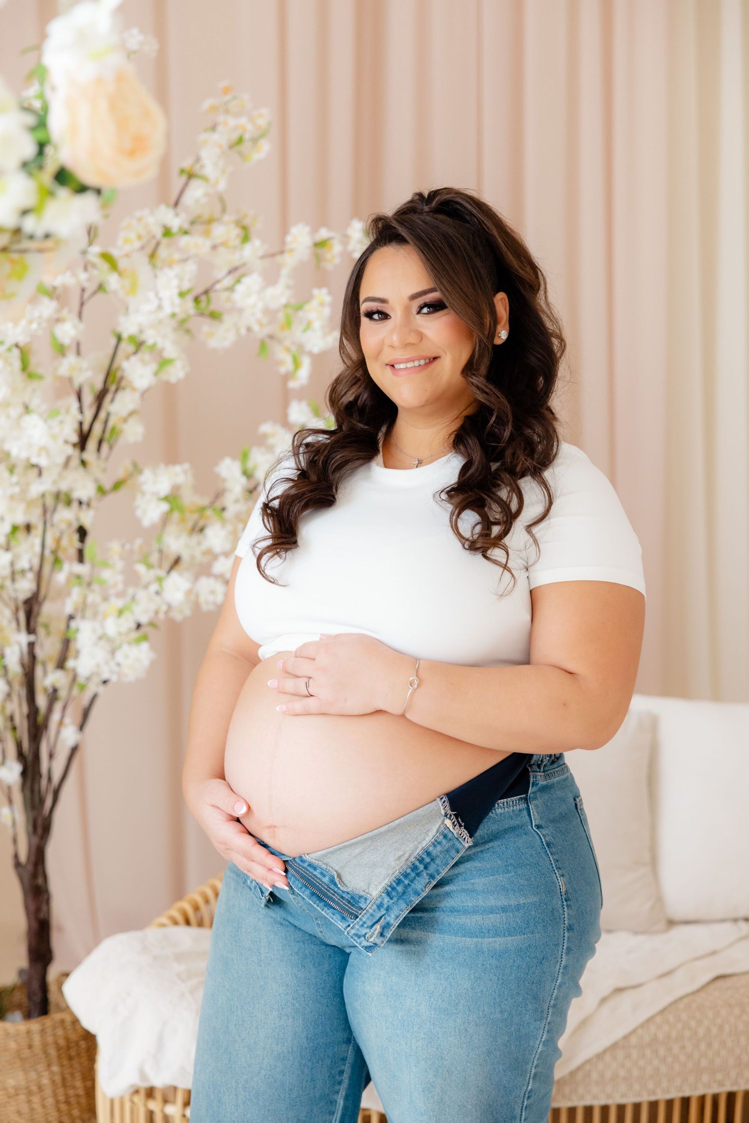 A pregnant woman in a white t-shirt and jeans poses by spring cherry blossoms in a feminine home studio setting.
