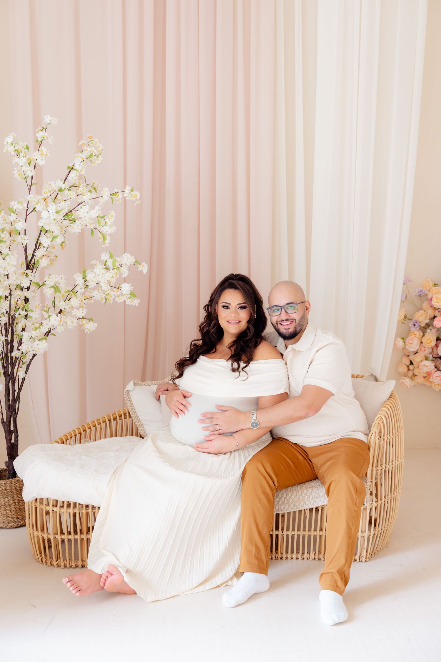 Couple sits together in romantic indoor setting with cherry blossoms and rattan furniture against pink curtained backdrop.