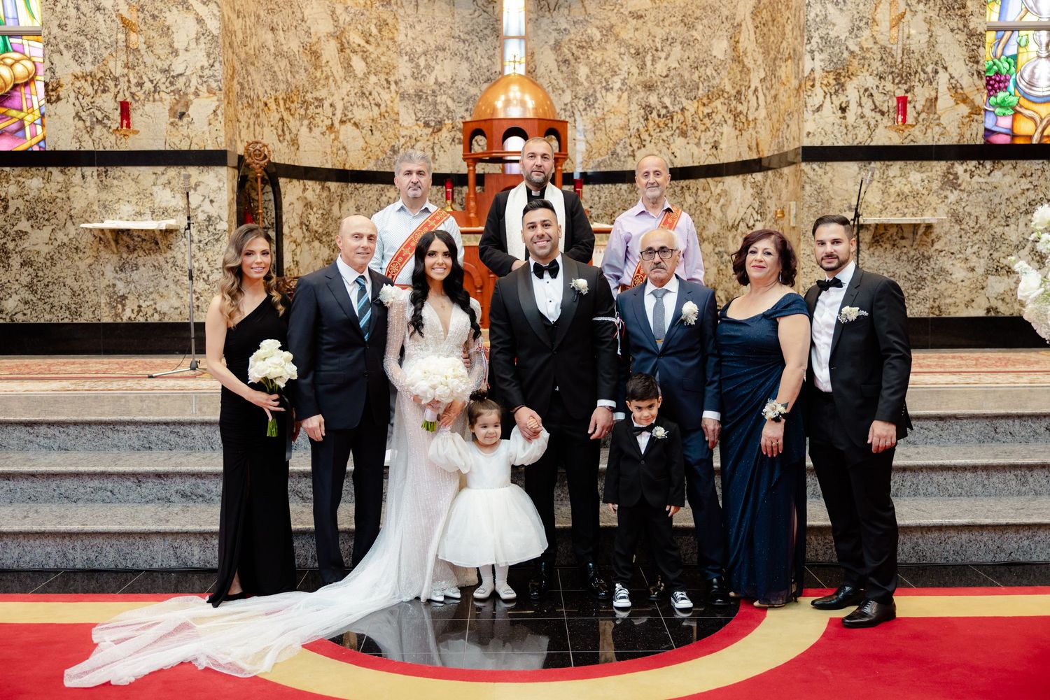 Wedding party gathers for formal photo on church steps with decorative cross and stained glass behind them.