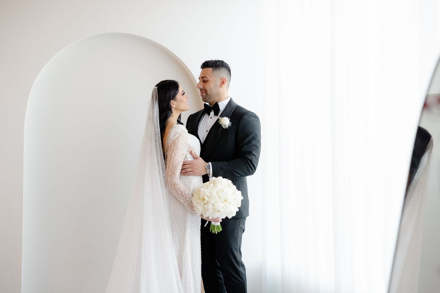 Intimate wedding portrait of couple in white archway with flowing veil and bouquet.