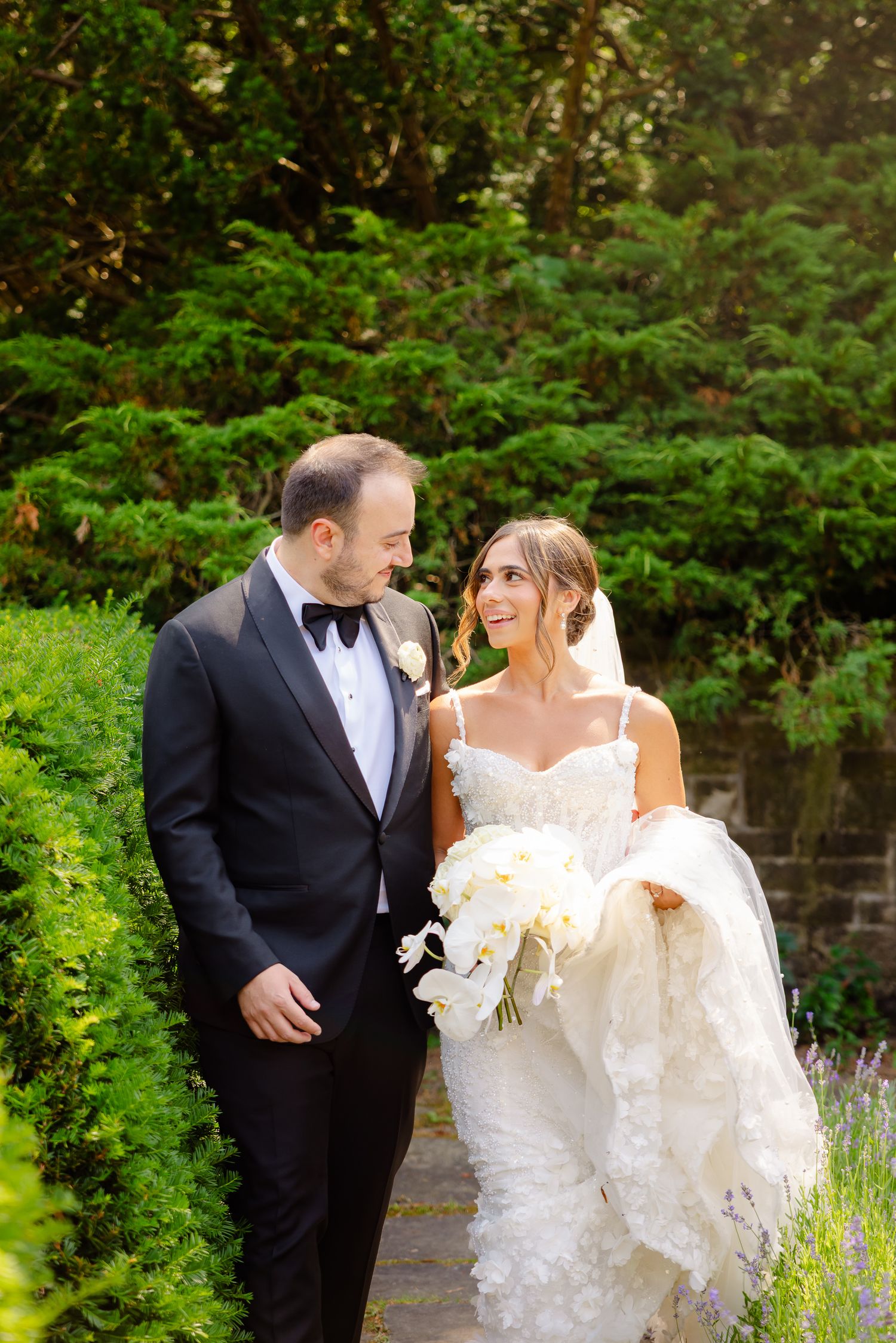 Loving couple in formal wedding attire stroll along garden path surrounded by lush greenery.