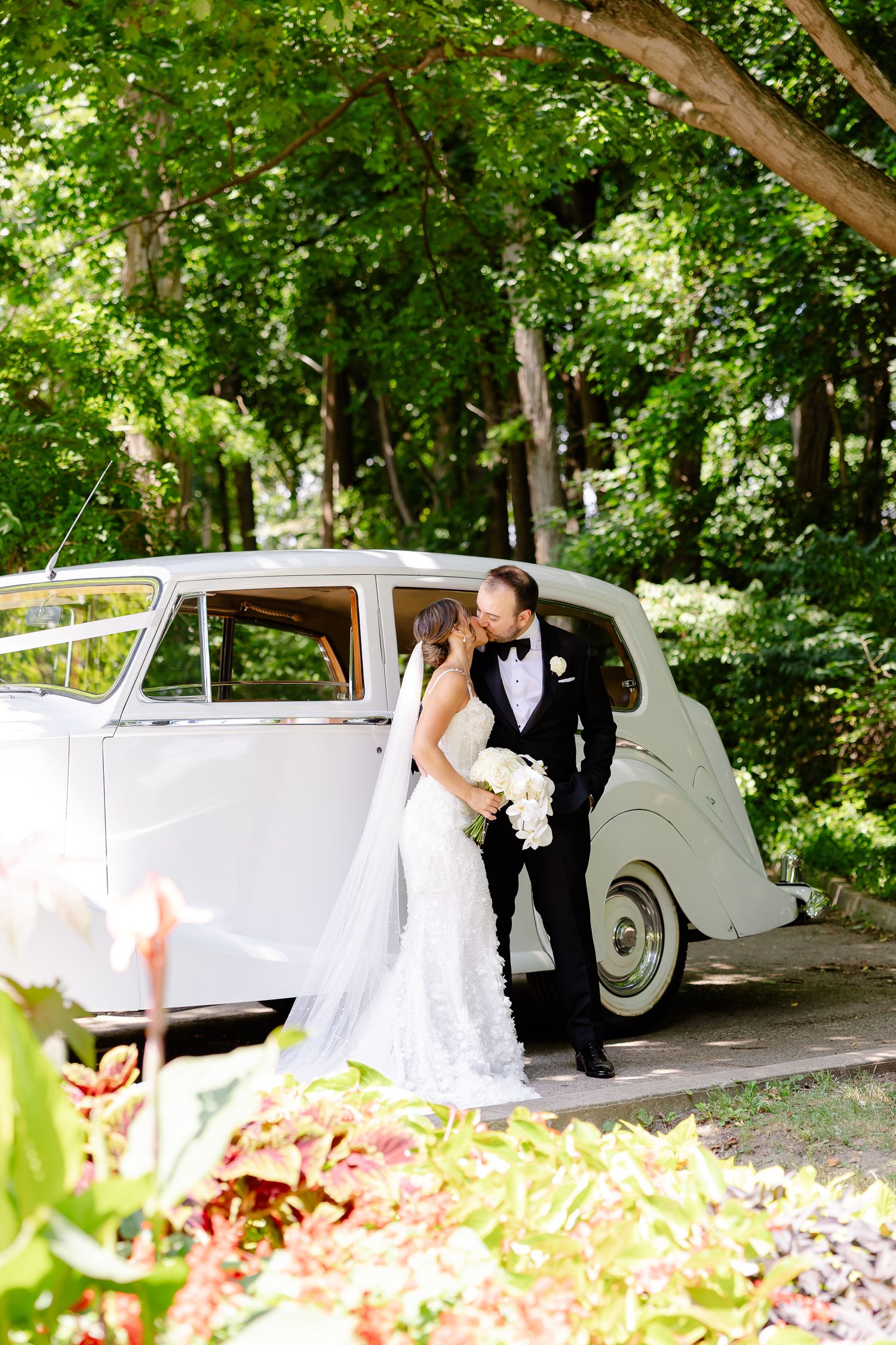 A classic white vintage car serves as an elegant backdrop for a couple on their wedding day in a lush garden setting.