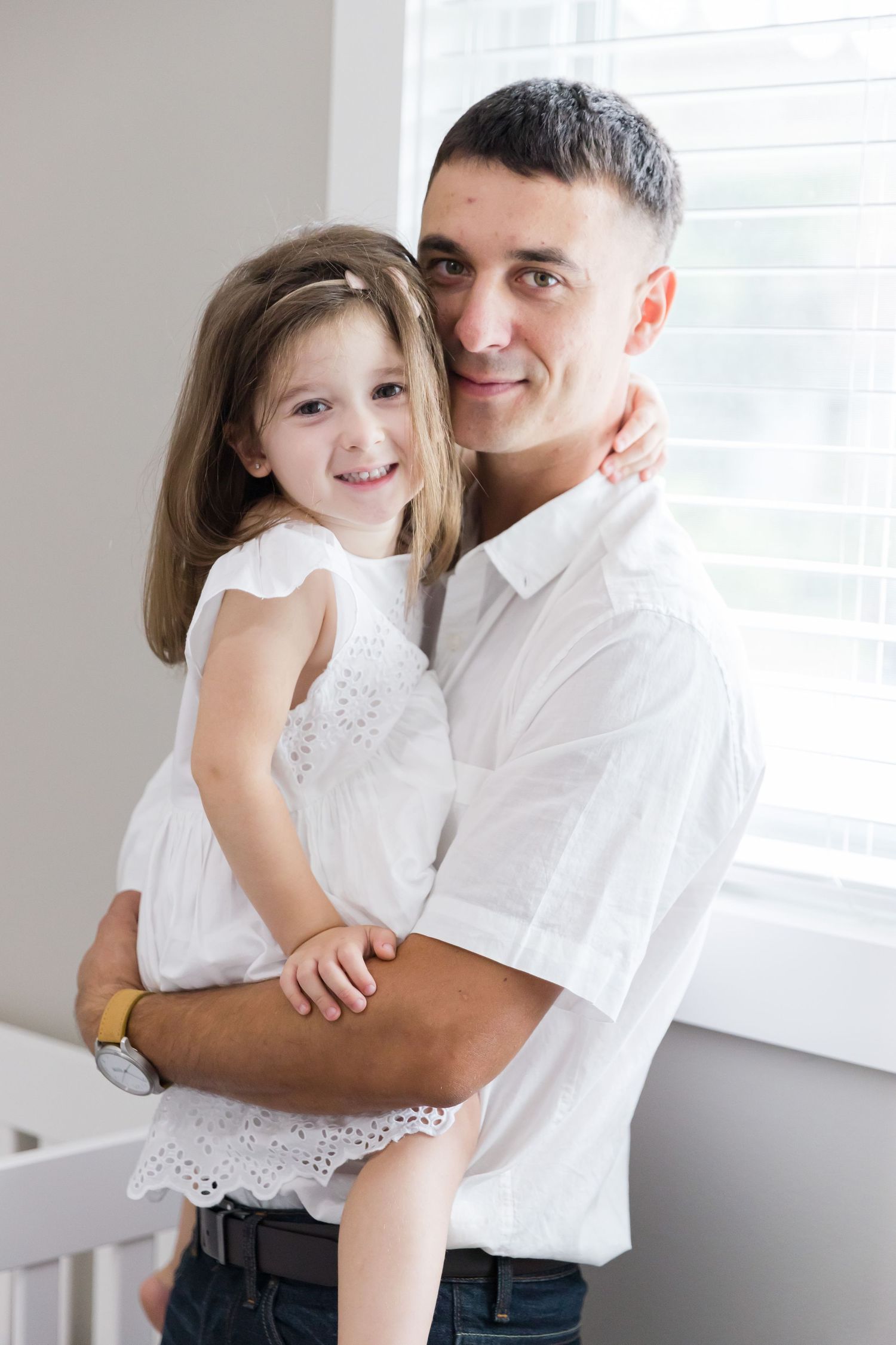 Parent and child in matching white outfits share joyful moment near window.