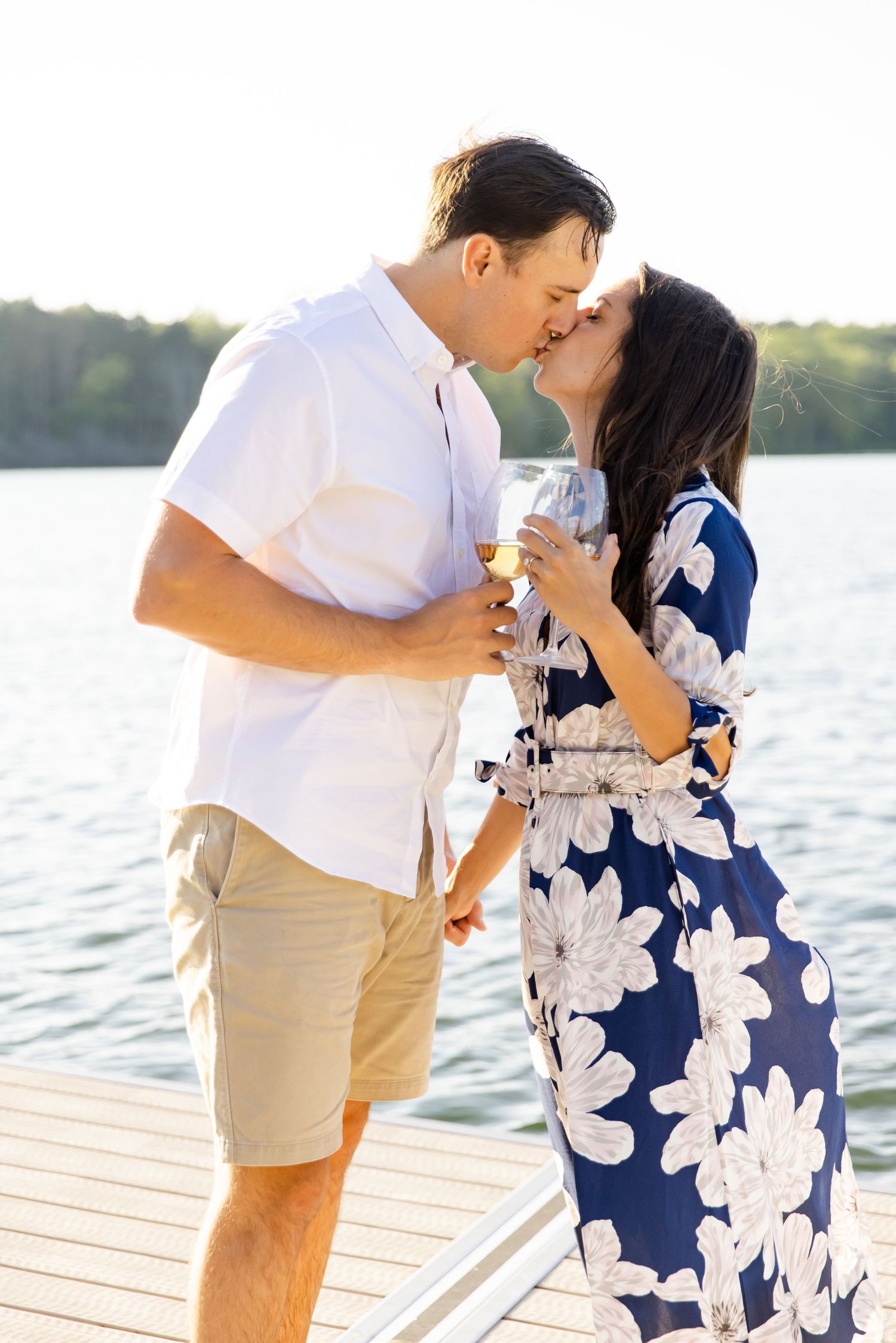 Couple sharing a romantic kiss while holding drinks on a lakeside dock at sunset.