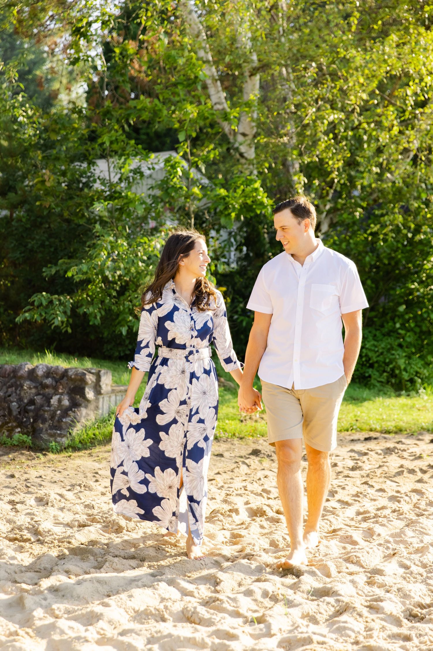 A couple walking barefoot on a sandy beach wearing a floral dress and white shirt against a green forest backdrop.