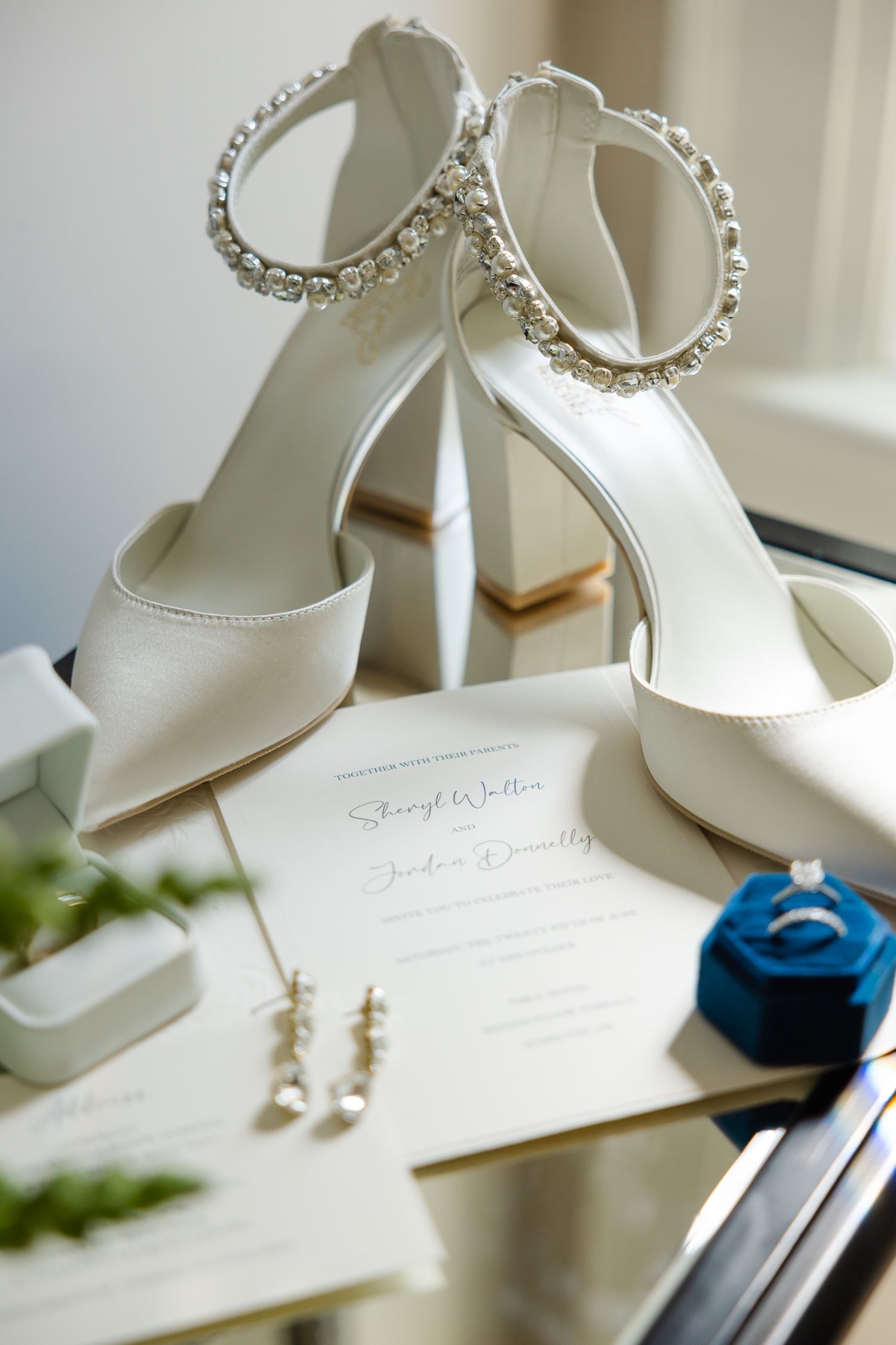 Elegant white bridal shoes with crystal details alongside wedding invitation and jewelry.