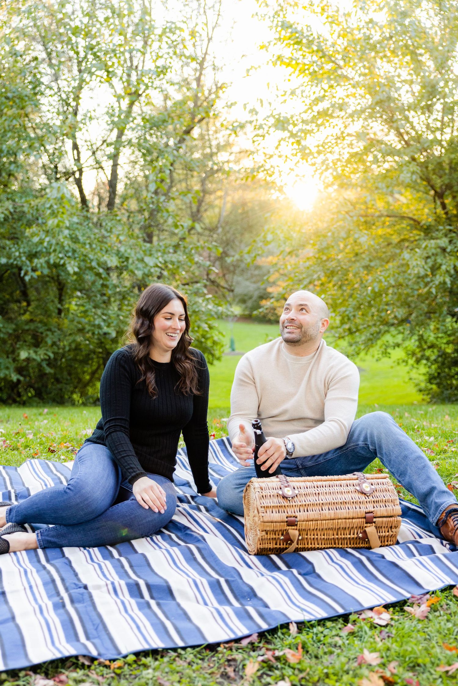 Couple relaxing on blue striped blanket with wicker basket in sunlit park setting.