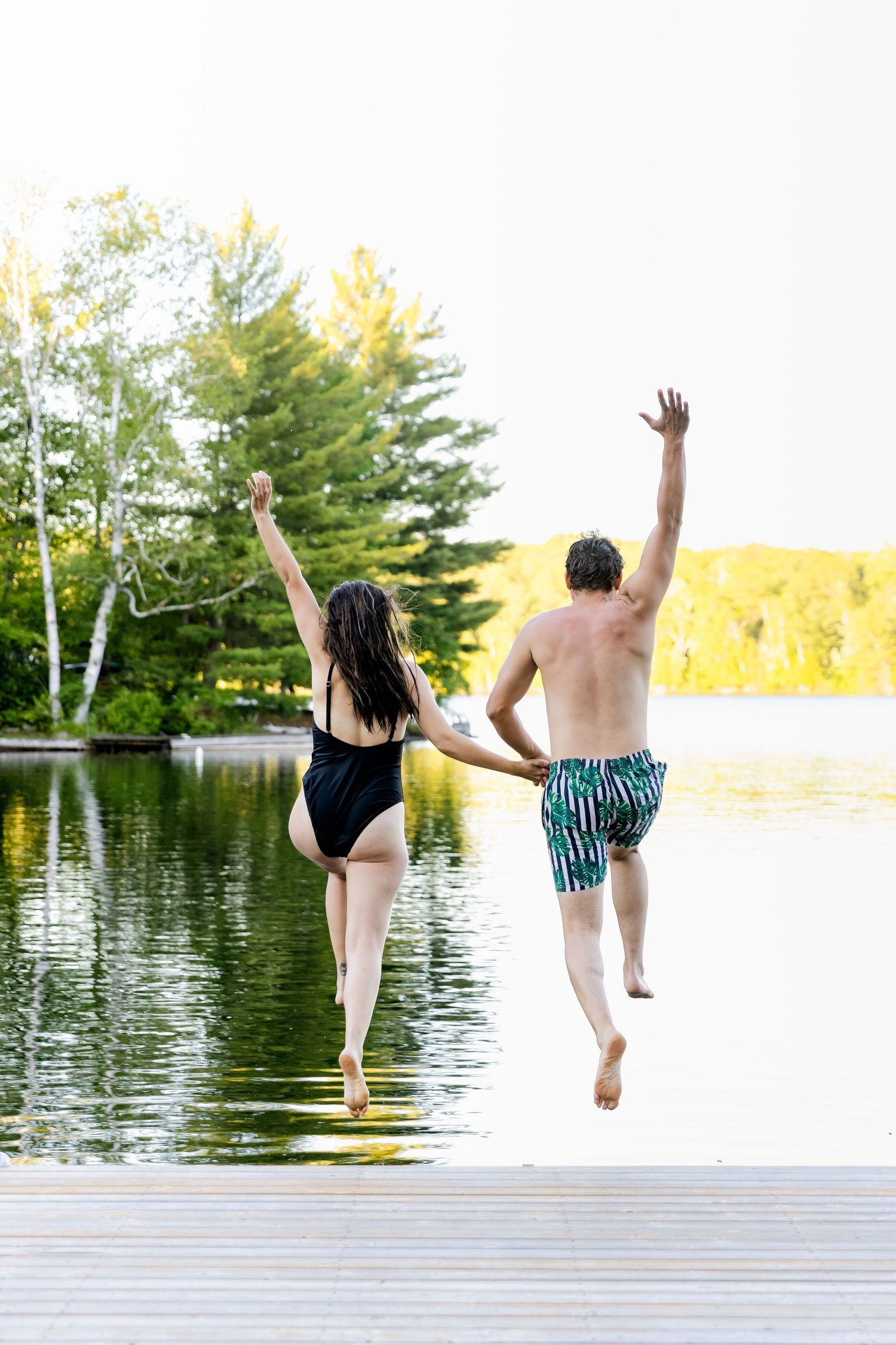 Two people jumping joyfully off a wooden dock into a lake during summer.