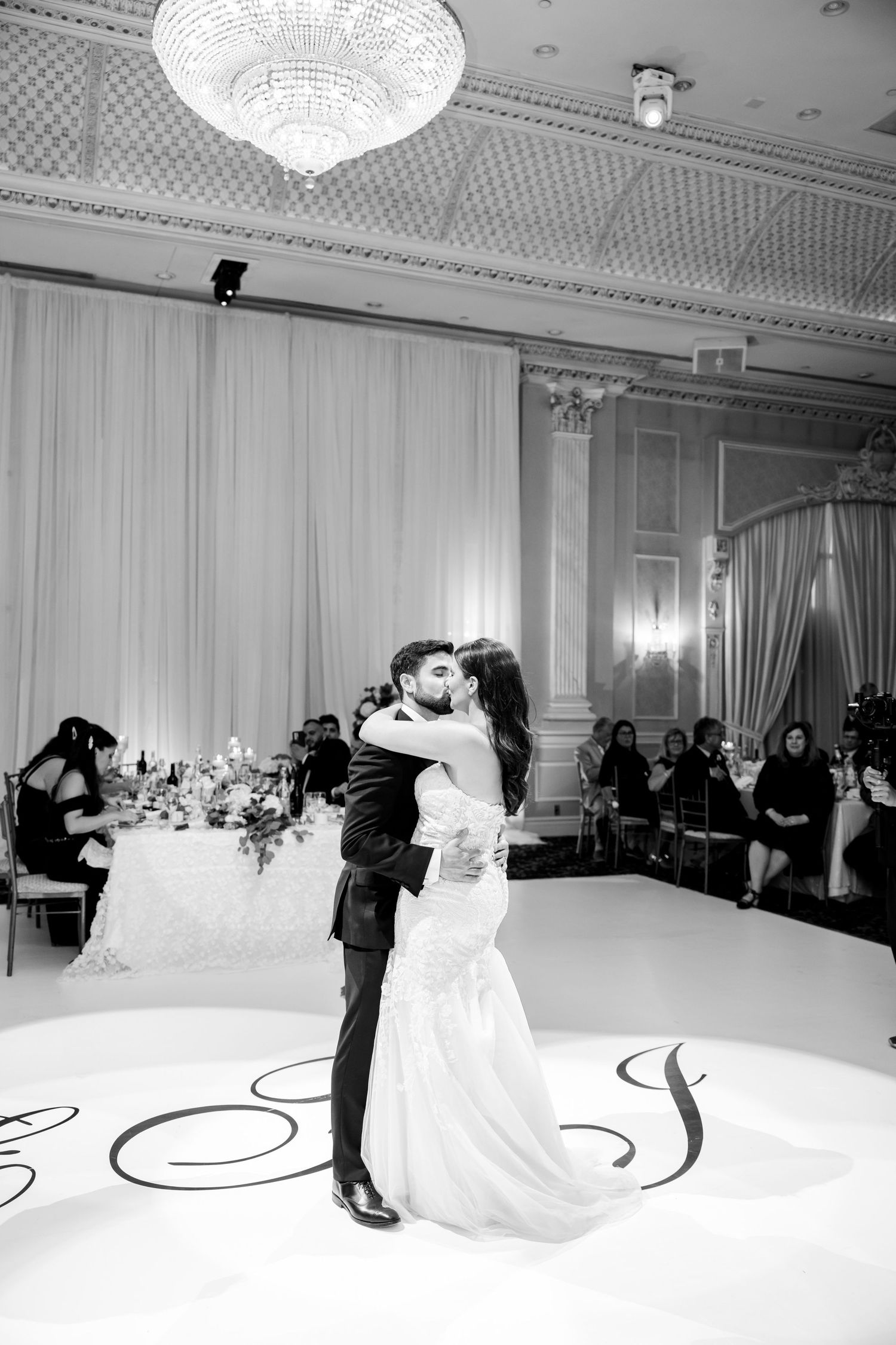 A romantic dance captured in black and white under a crystal chandelier with guests looking on.