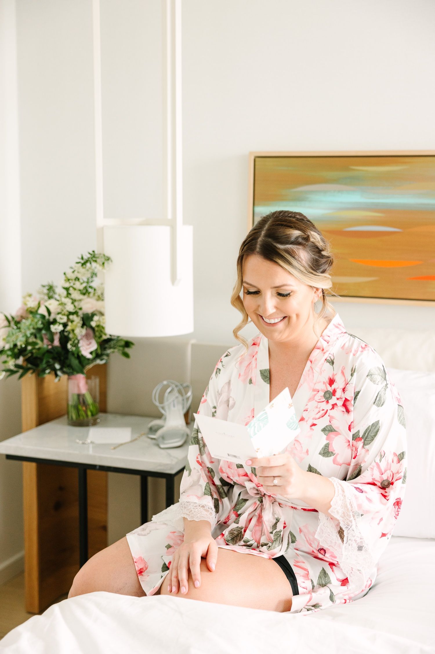 Bride in floral robe sitting on bed with abstract art and flowers in background.