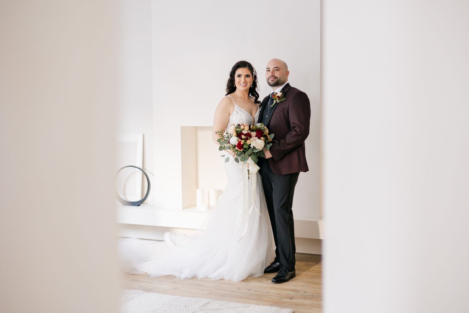 Newlywed couple in formal wedding attire stand together against white wall.
