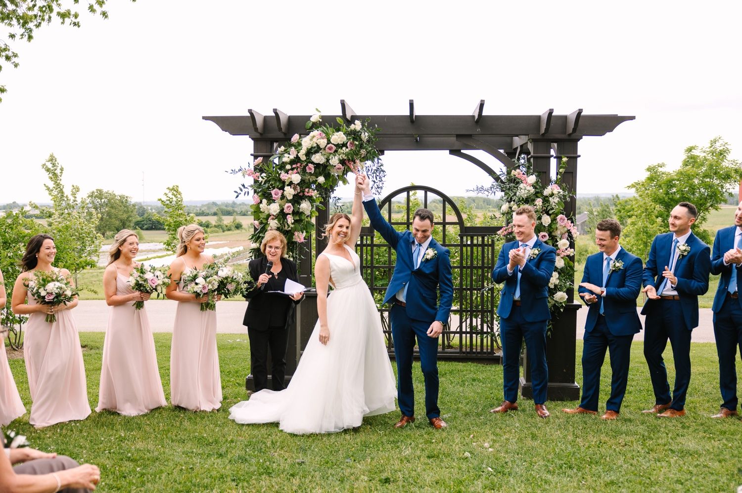Wedding party poses under decorated pergola with bridesmaids in pink and groomsmen in blue.