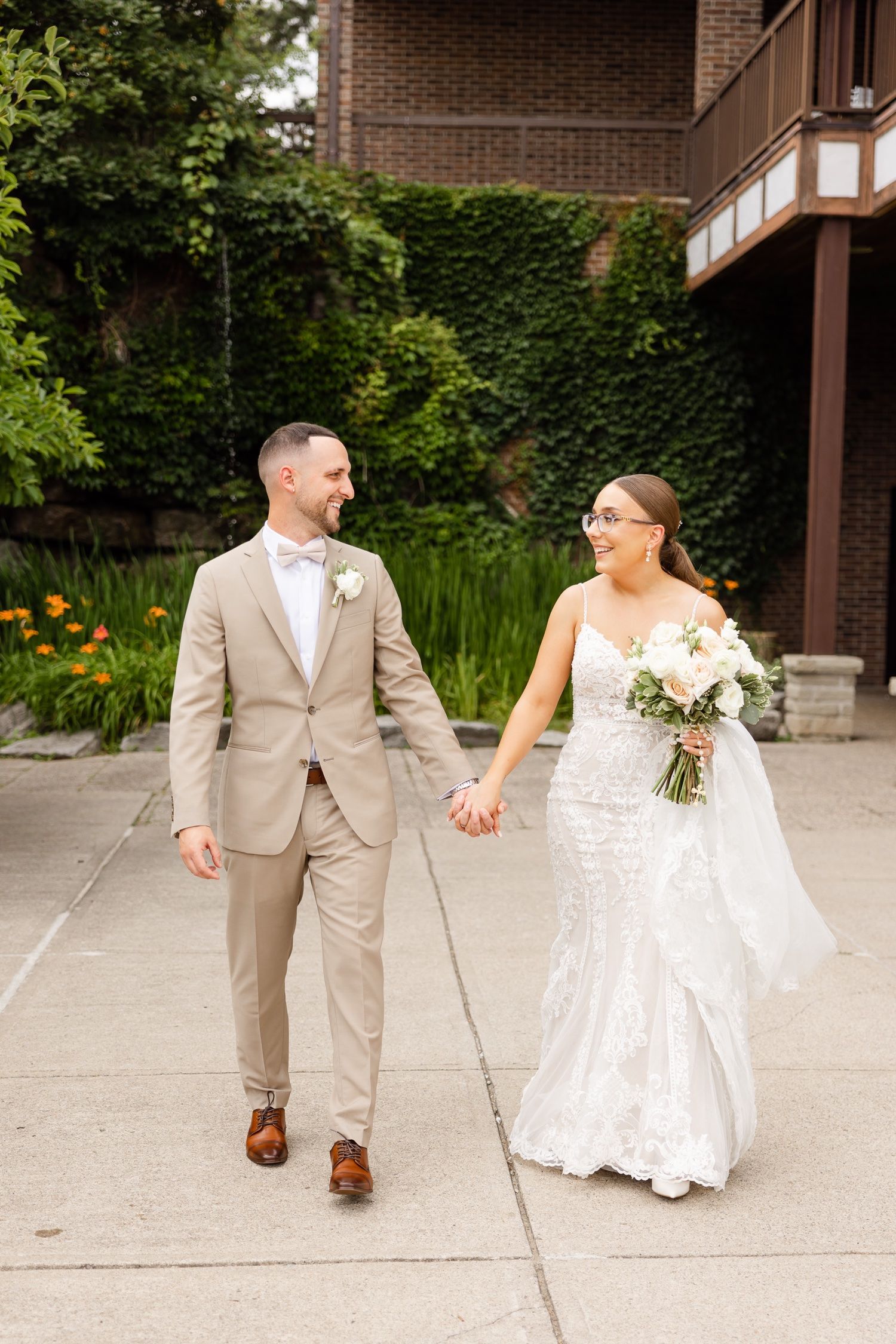 Newlyweds walk hand in hand along garden pathway in tan suit and lace gown.