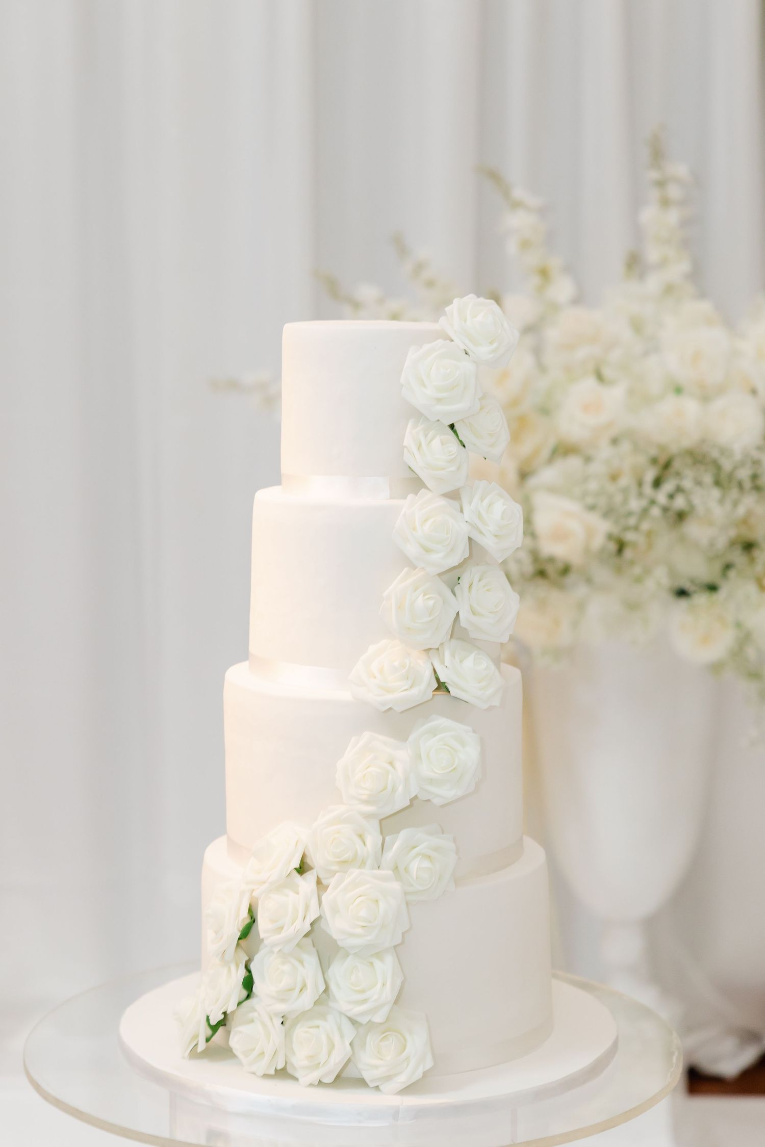 Four-tier white wedding cake decorated with cascading white roses against a light backdrop.