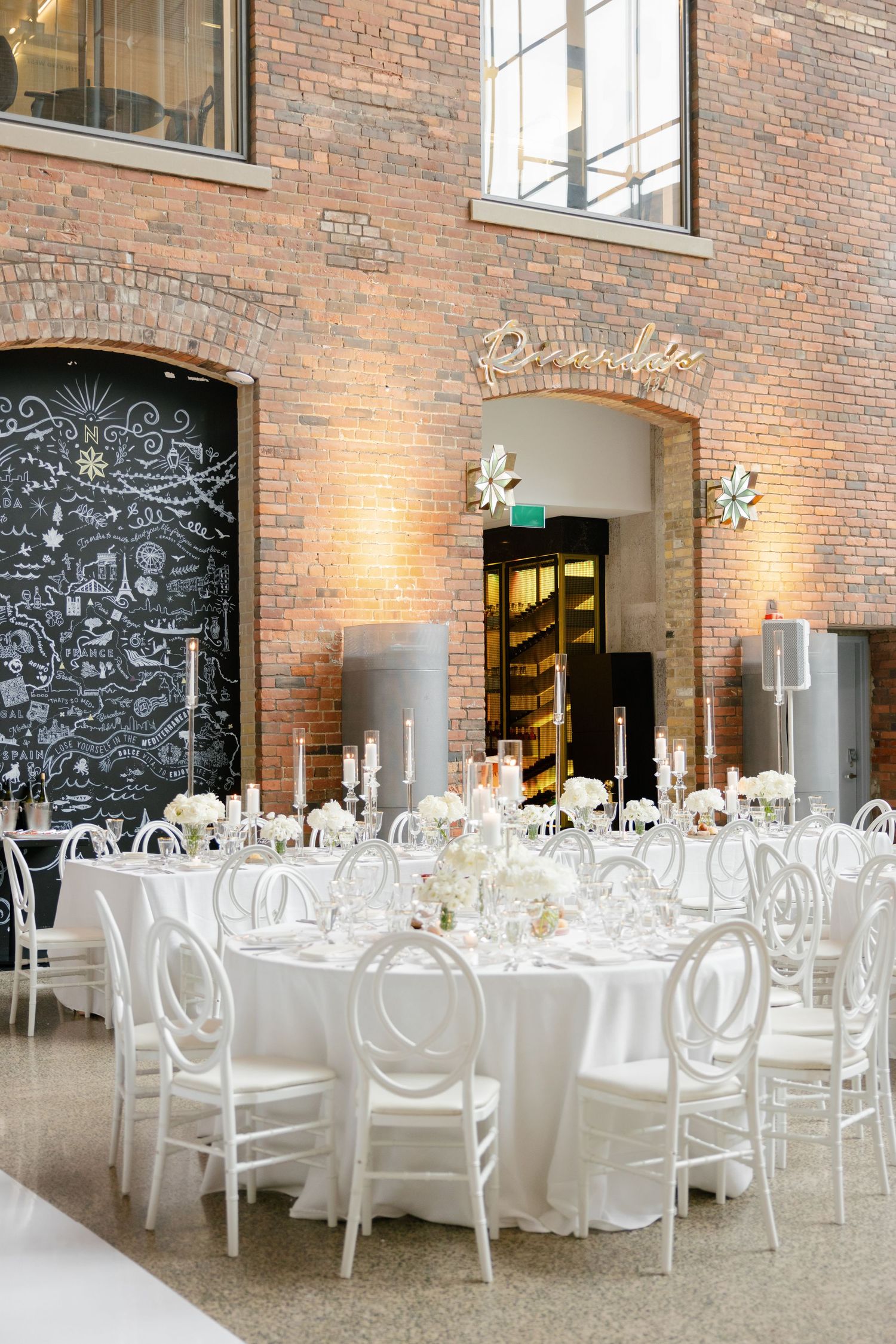 Elegant wedding reception setup with white tables and chairs in a rustic brick venue with chalkboard wall decor.