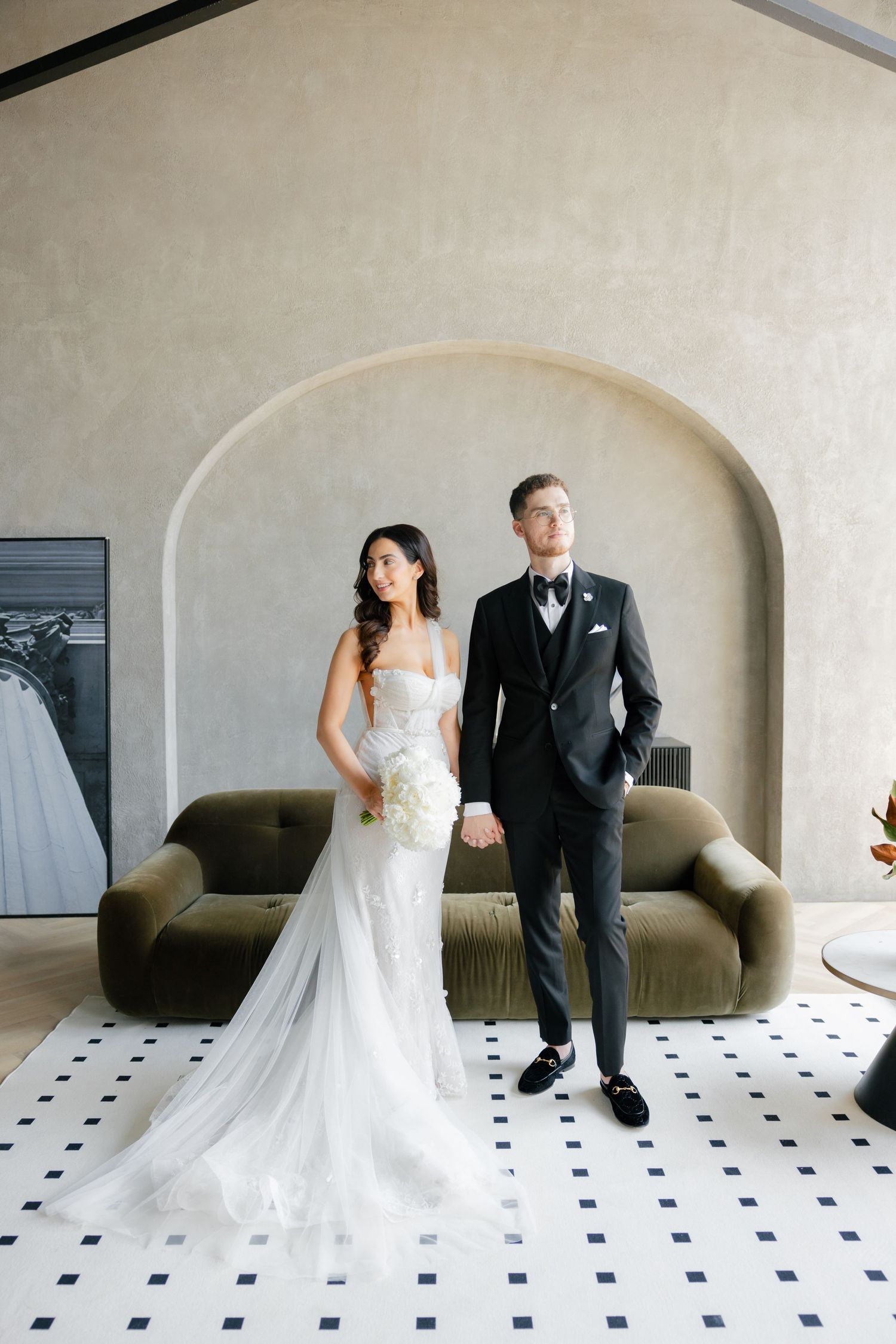 Elegant couple poses in wedding attire against arched wall with olive velvet sofa and geometric tile floor.
