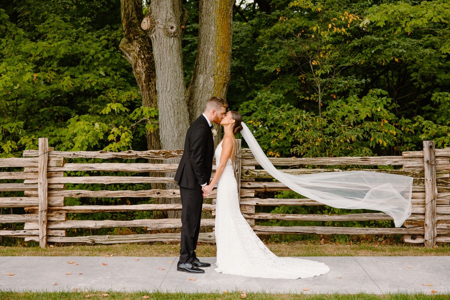 A romantic wedding moment captured in front of wooden fencing with a flowing veil in the breeze.