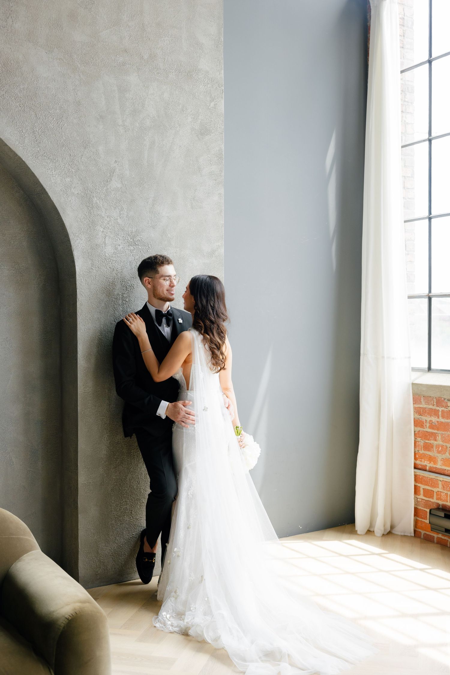 Romantic wedding couple poses against grey wall and large window in modern industrial space.