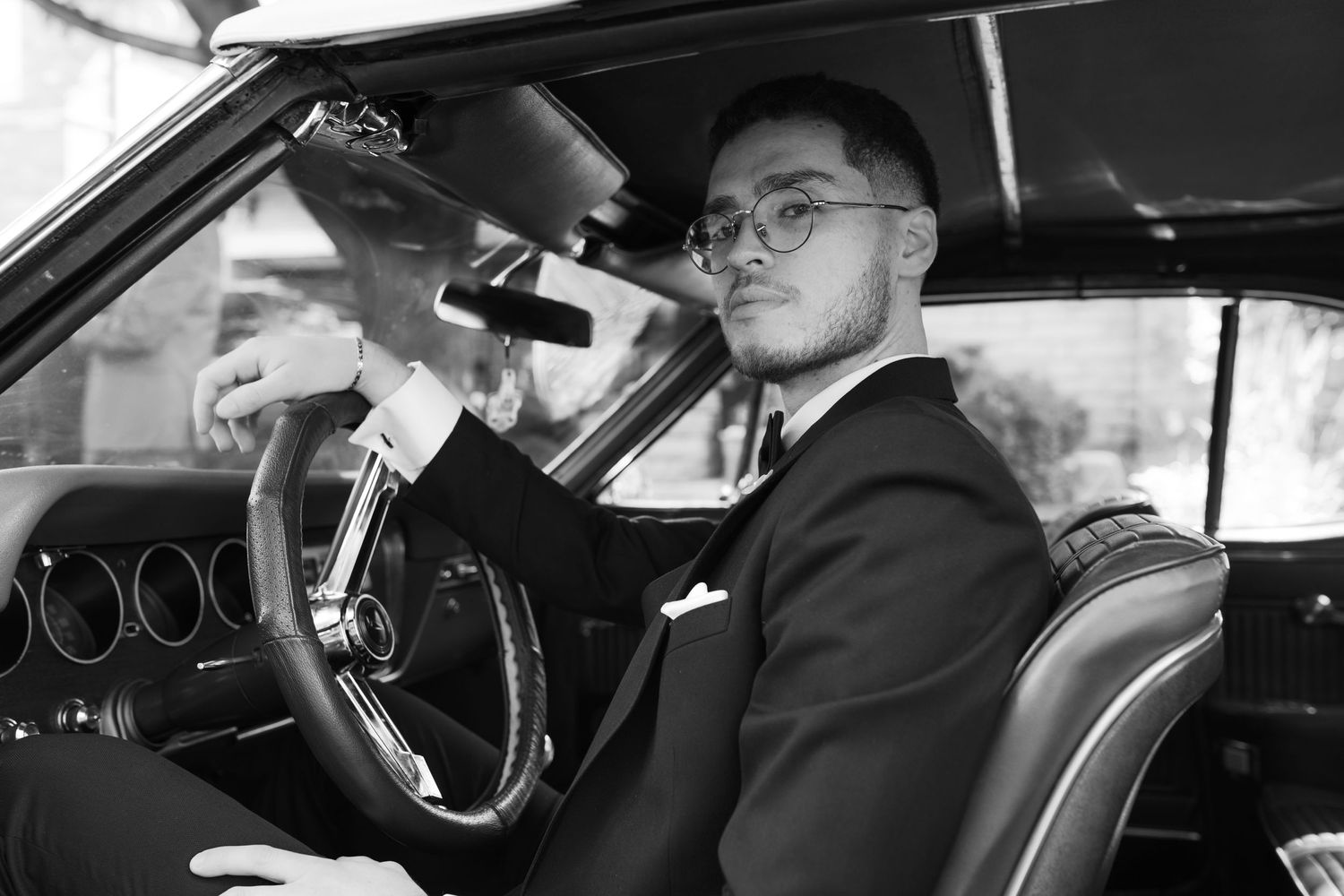Black and white photo of person in suit adjusting rearview mirror inside vintage car.