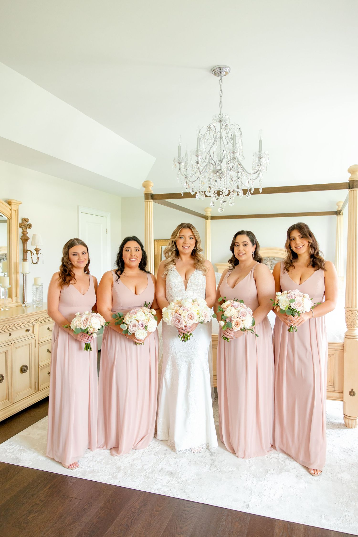 Wedding party in blush pink bridesmaid dresses and white gown posing under crystal chandelier.